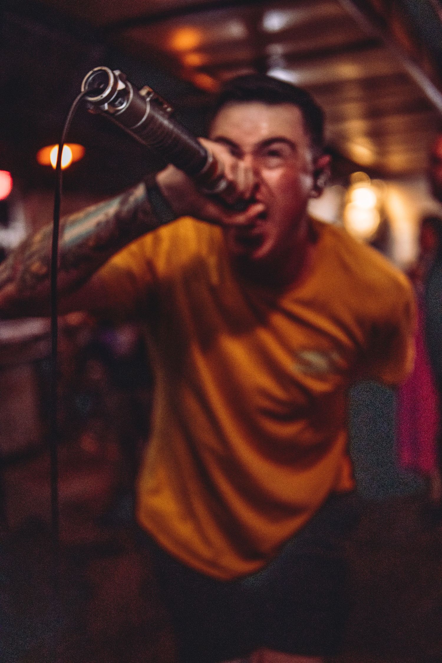 Blurred action shot of tattooed singer in yellow shirt screaming into mic at energetic punk concert.