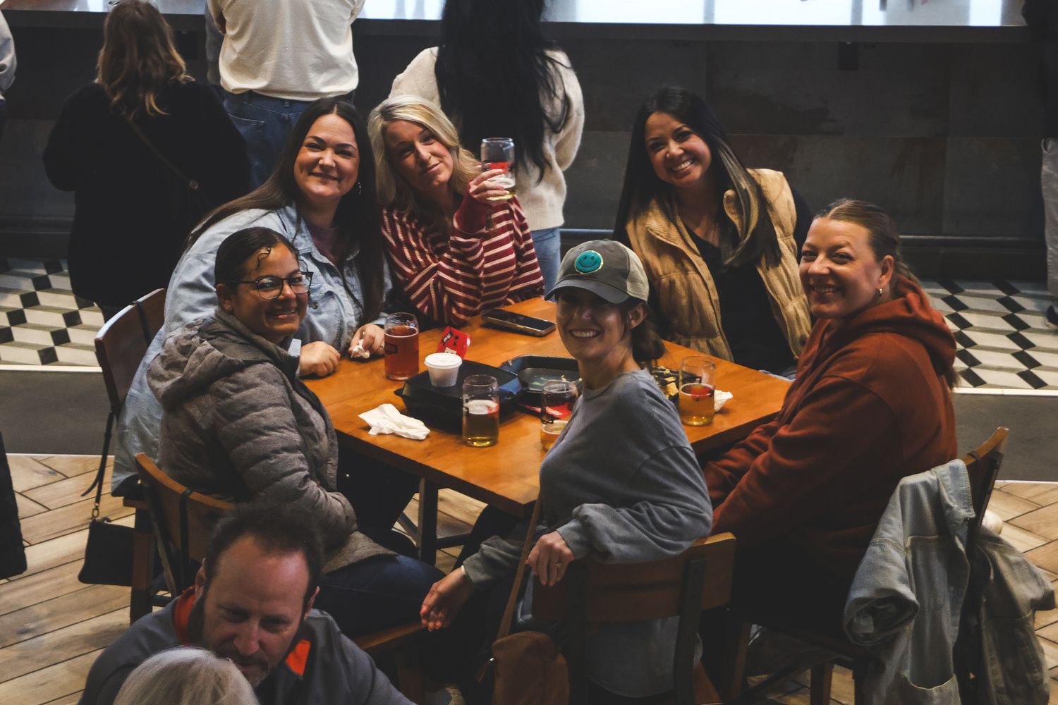 Group of friends smiling and gathered around a table at a casual indoor brewery or bar setting.