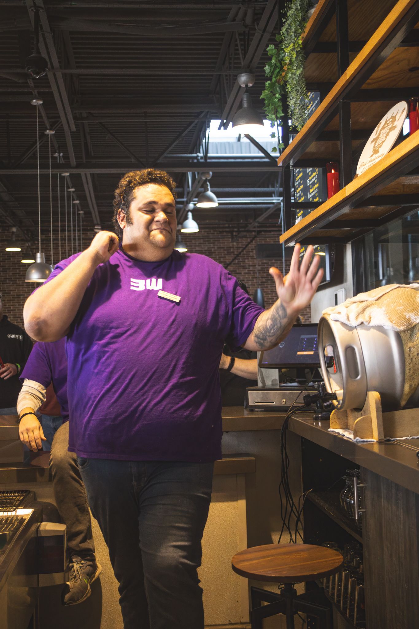 Man in purple brewery shirt waving near a keg at a bar counter.