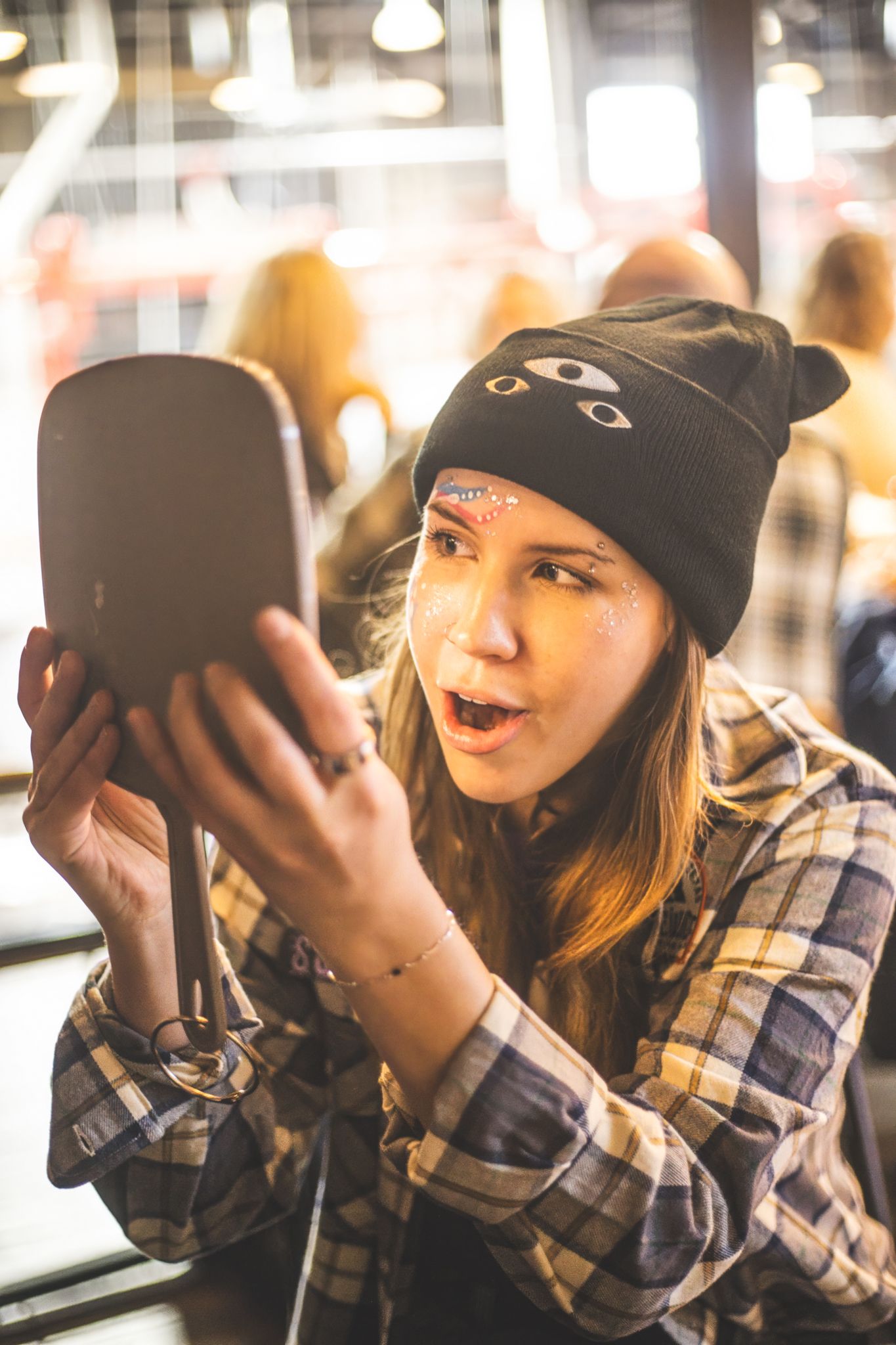 Young woman in plaid shirt and black beanie looking surprised at a handheld mirror indoors.