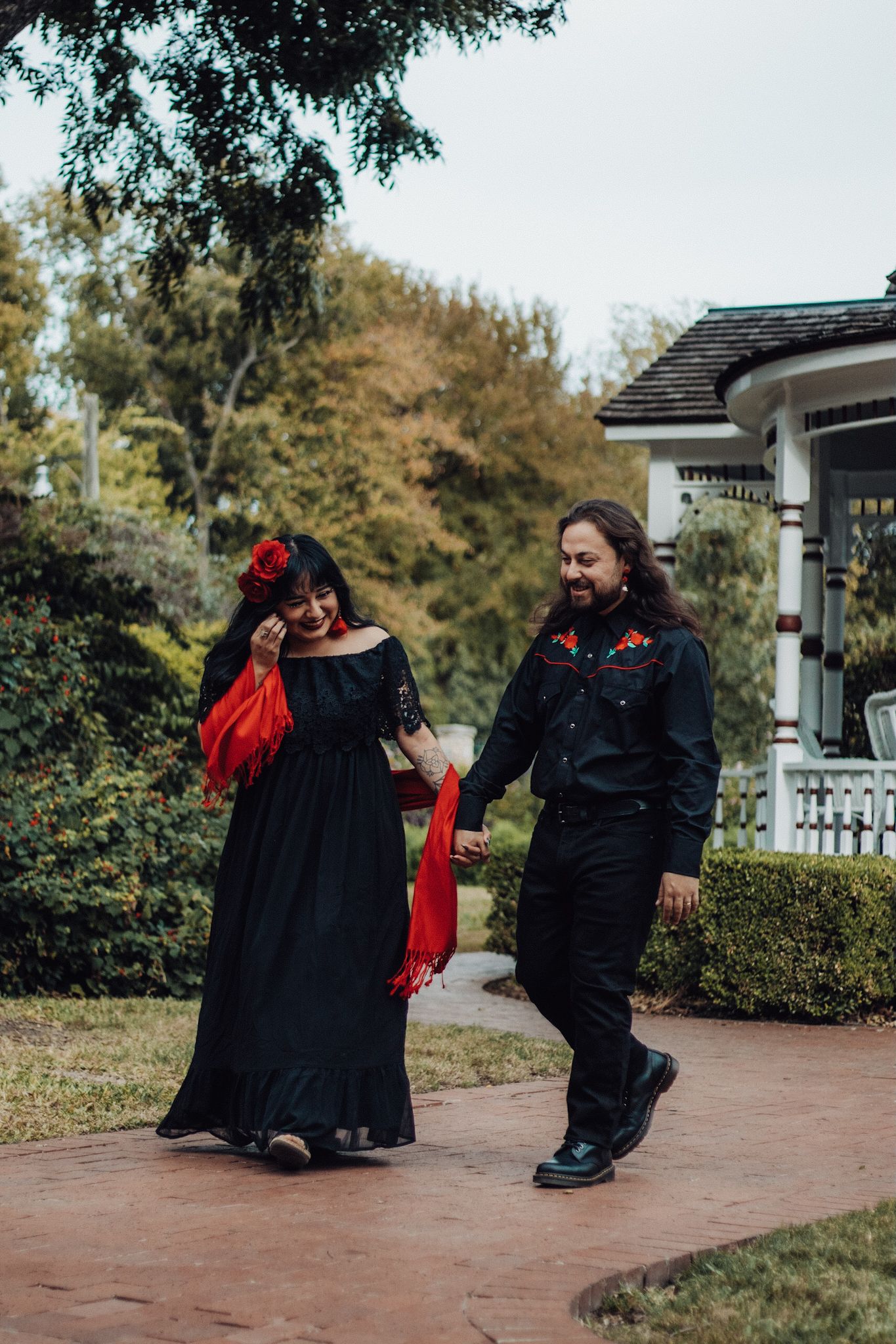 Couple in matching black outfits with red accents holding hands while walking on a garden path near a white house.