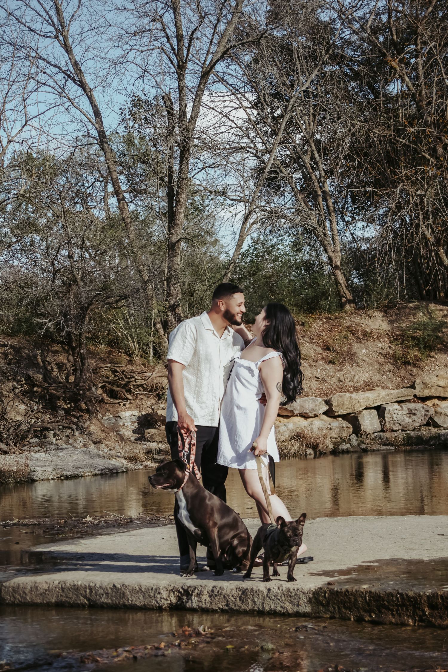 Couple in white outfits standing with two dogs by a rocky creek surrounded by bare winter trees.