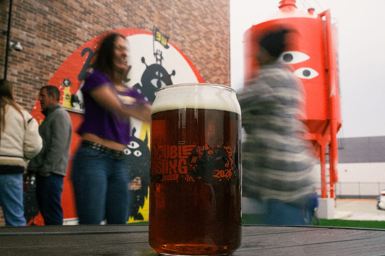 Close-up of a craft beer pint glass at a lively brewery with a red tank and dancing guests in the background.