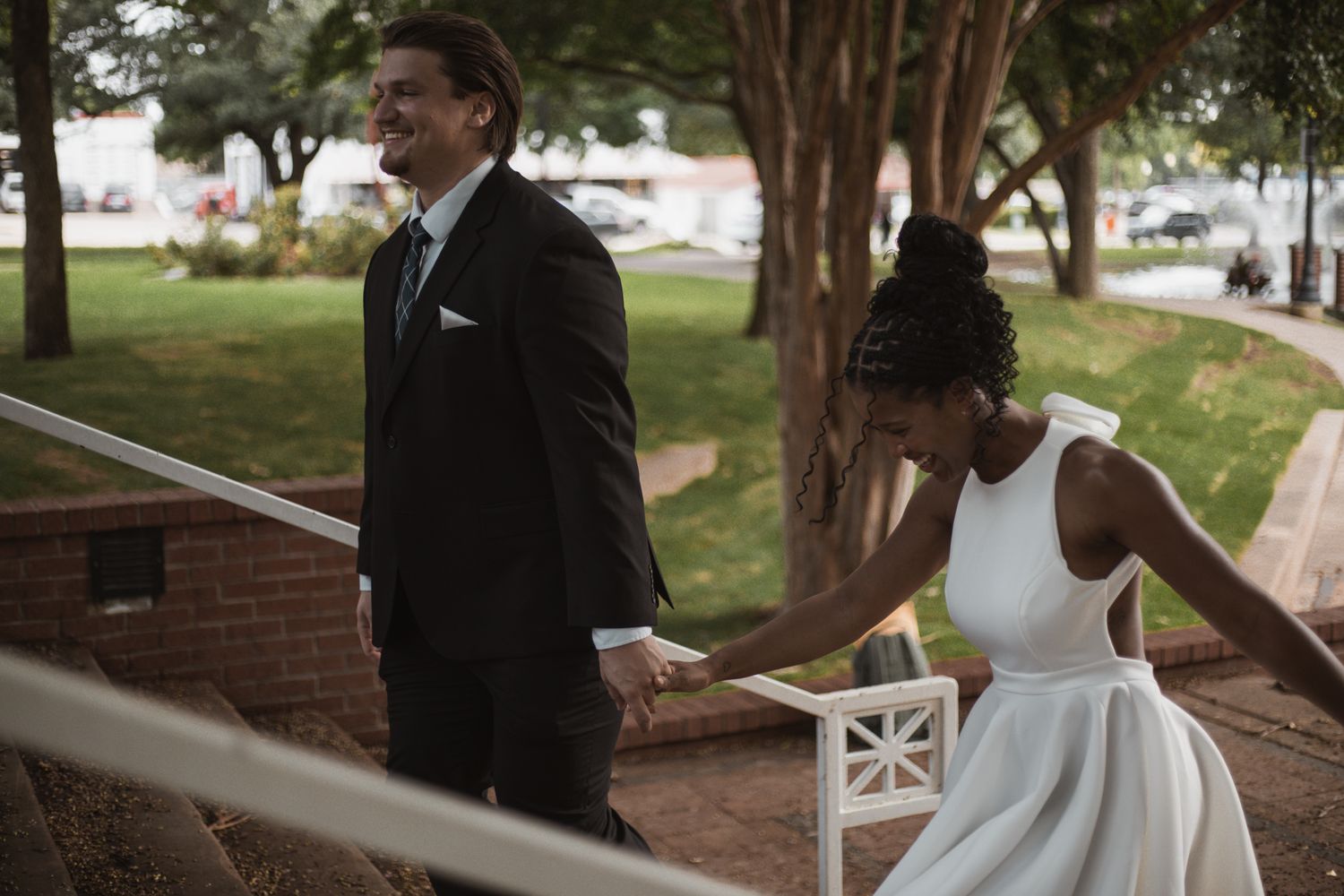 Interracial couple holding hands on outdoor stairs, groom in black suit and bride in white wedding dress.