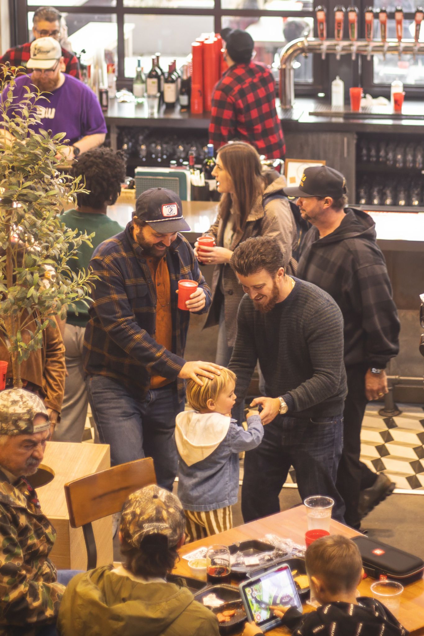 Crowd of people socializing inside a brewery taproom, with a child interacting with adults near a bar.
