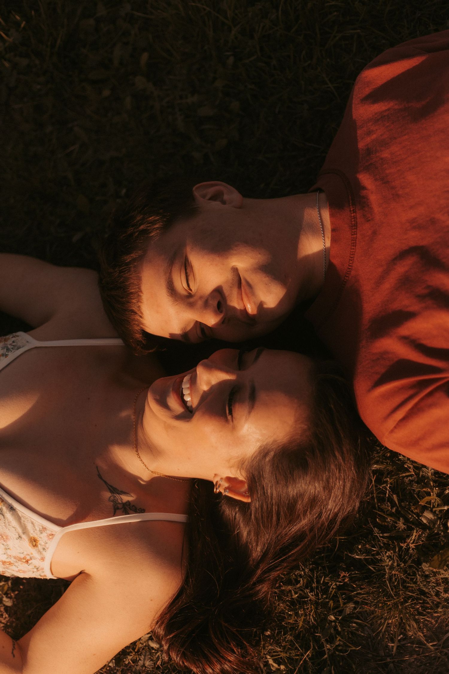 A happy couple lying on grass together, smiling and gazing at each other in warm golden sunlight.