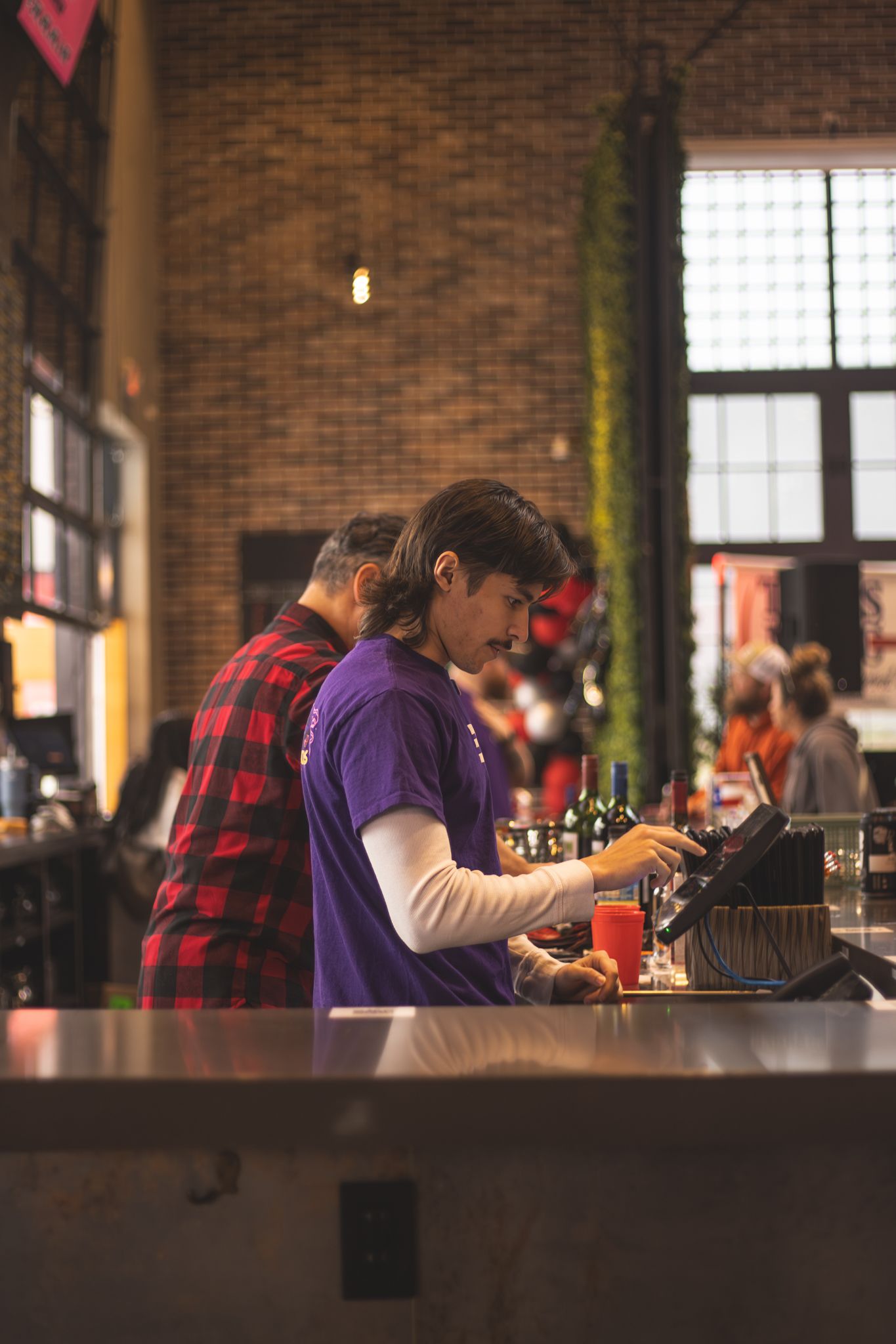 Bartender in purple shirt works at a rustic brick bar, processing orders on a tablet with drinks nearby.