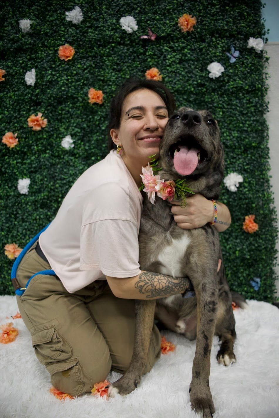 Smiling woman hugging a gray dog with a pink flower collar against a floral green wall.