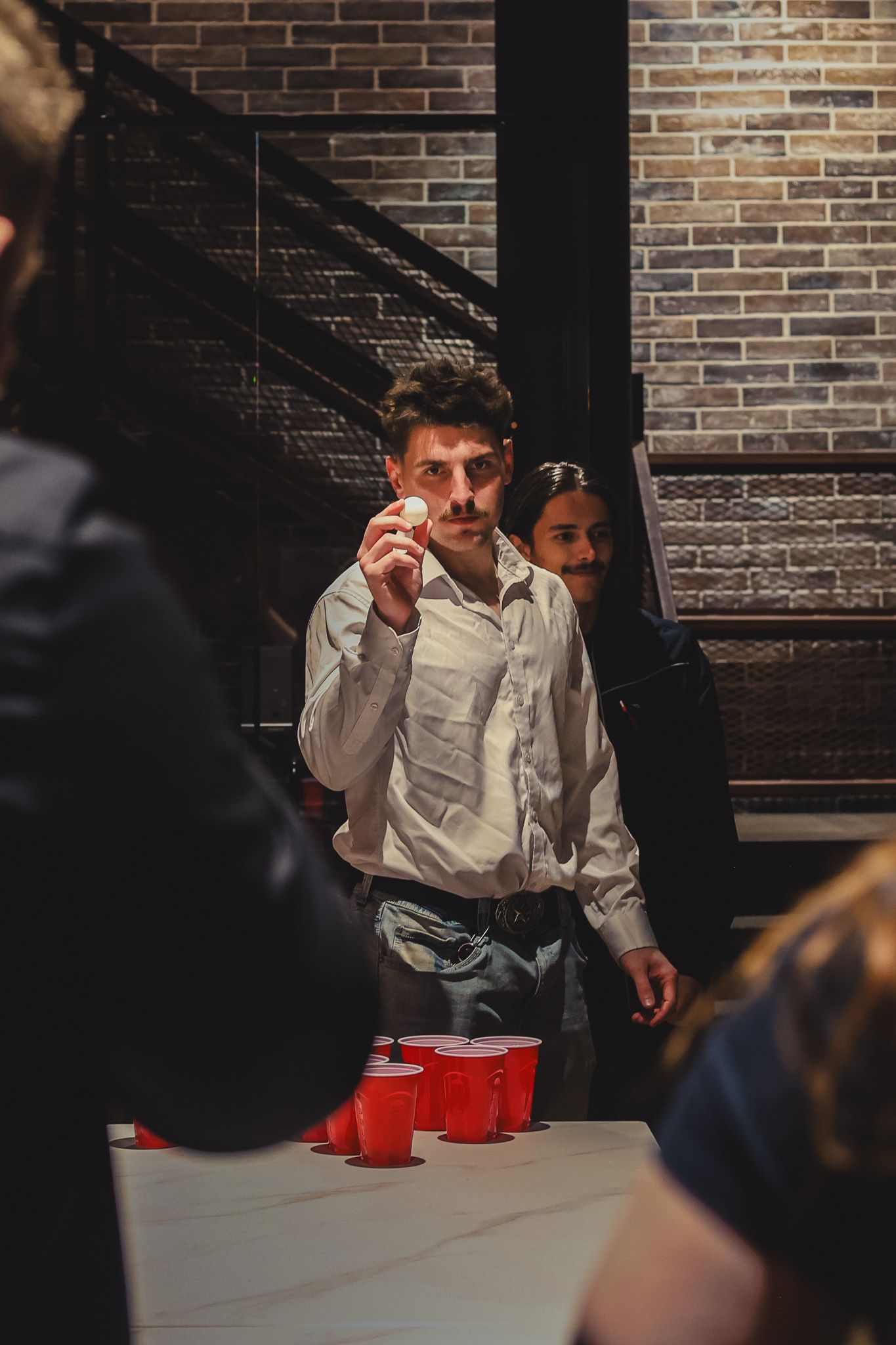 A young man prepares to throw a ping pong ball at red cups during a beer pong game.