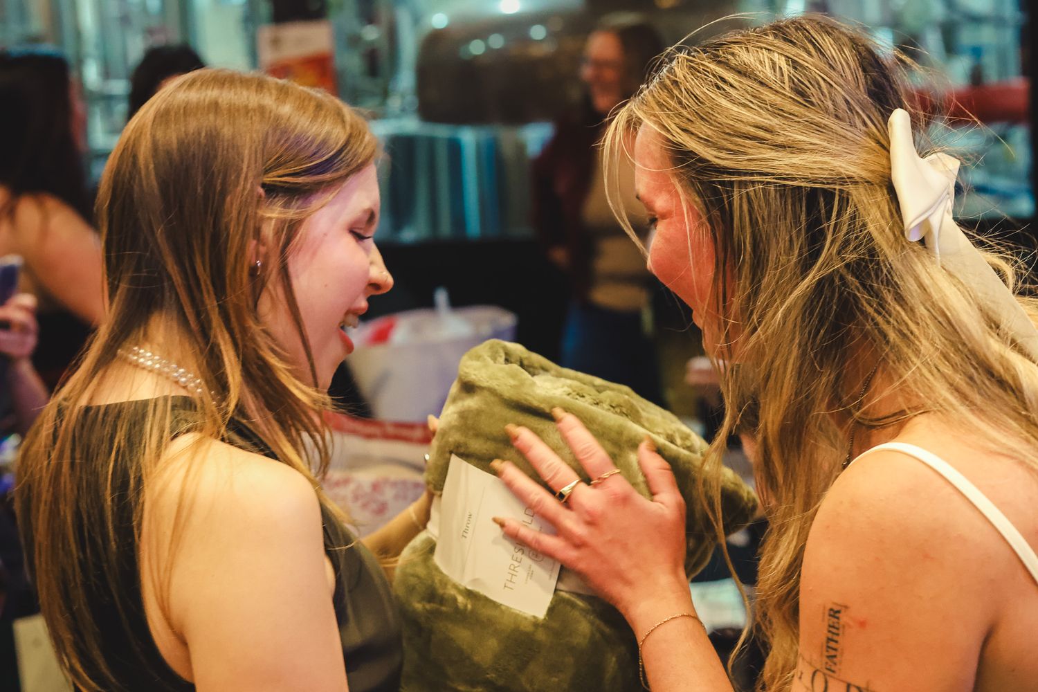 Two women with long hair laughing and sharing a green gift at an indoor market event.