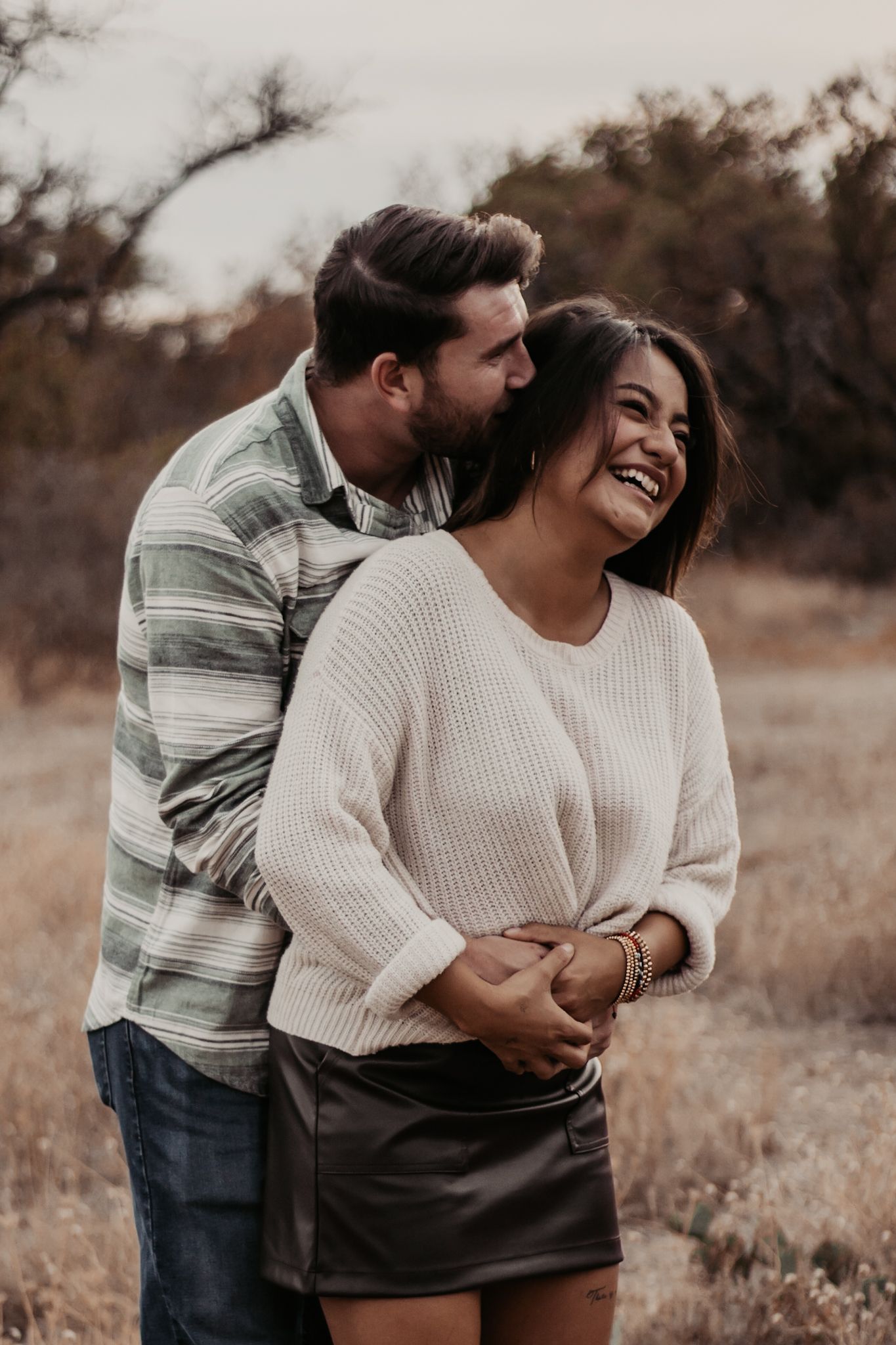 A couple embraces outdoors, woman laughing joyfully in white sweater and leather skirt, man in plaid shirt hugging her from behind.