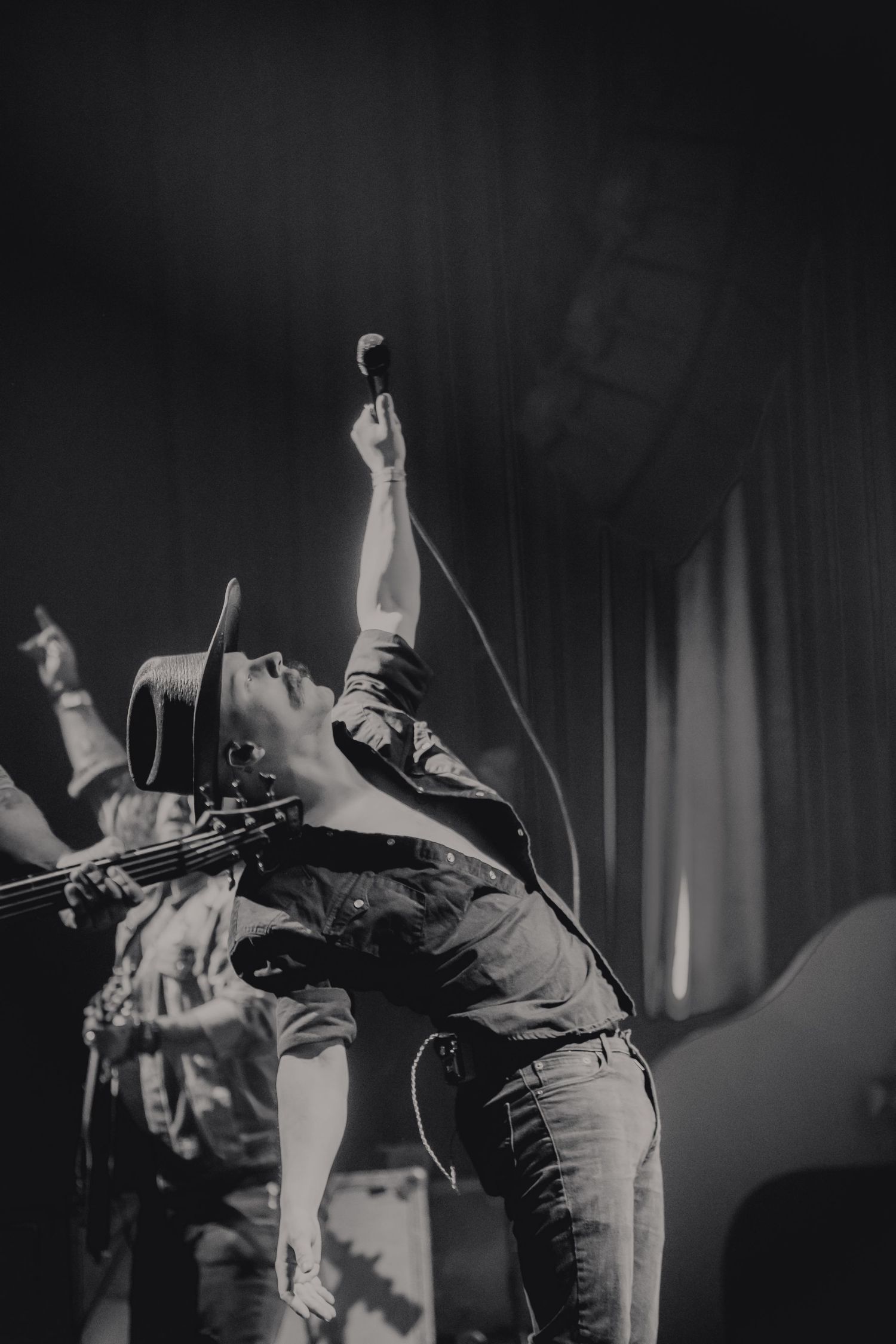 Black and white photo of a dynamic performer in a cowboy hat raising a microphone on stage.
