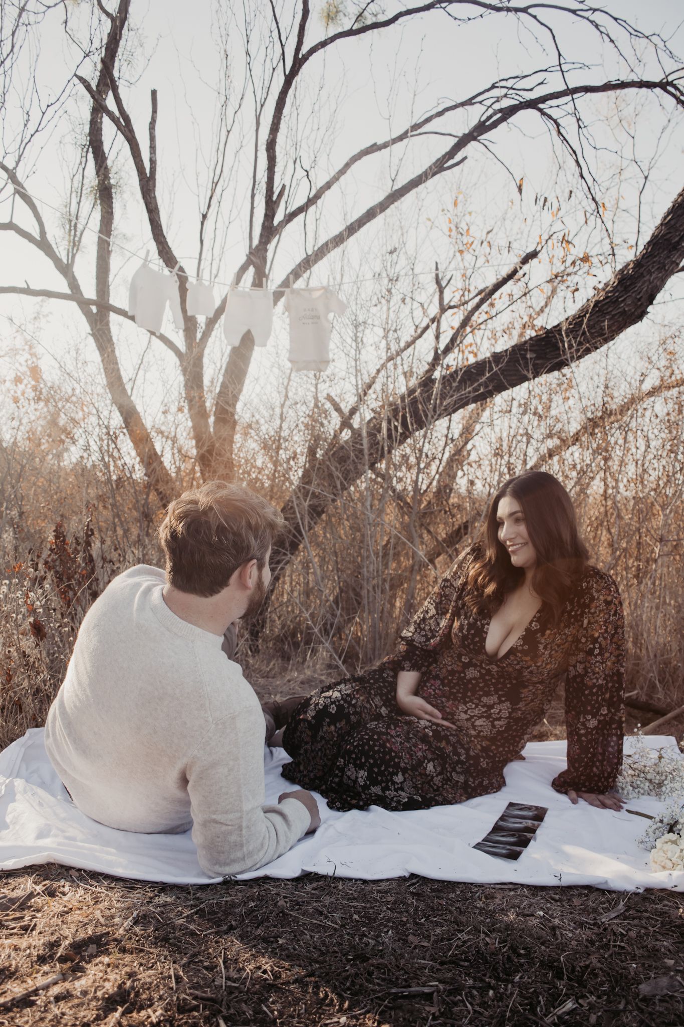 Couple sitting on white blanket outdoors near bare trees with hanging lanterns, woman in floral dress holding baby ultrasound photos.