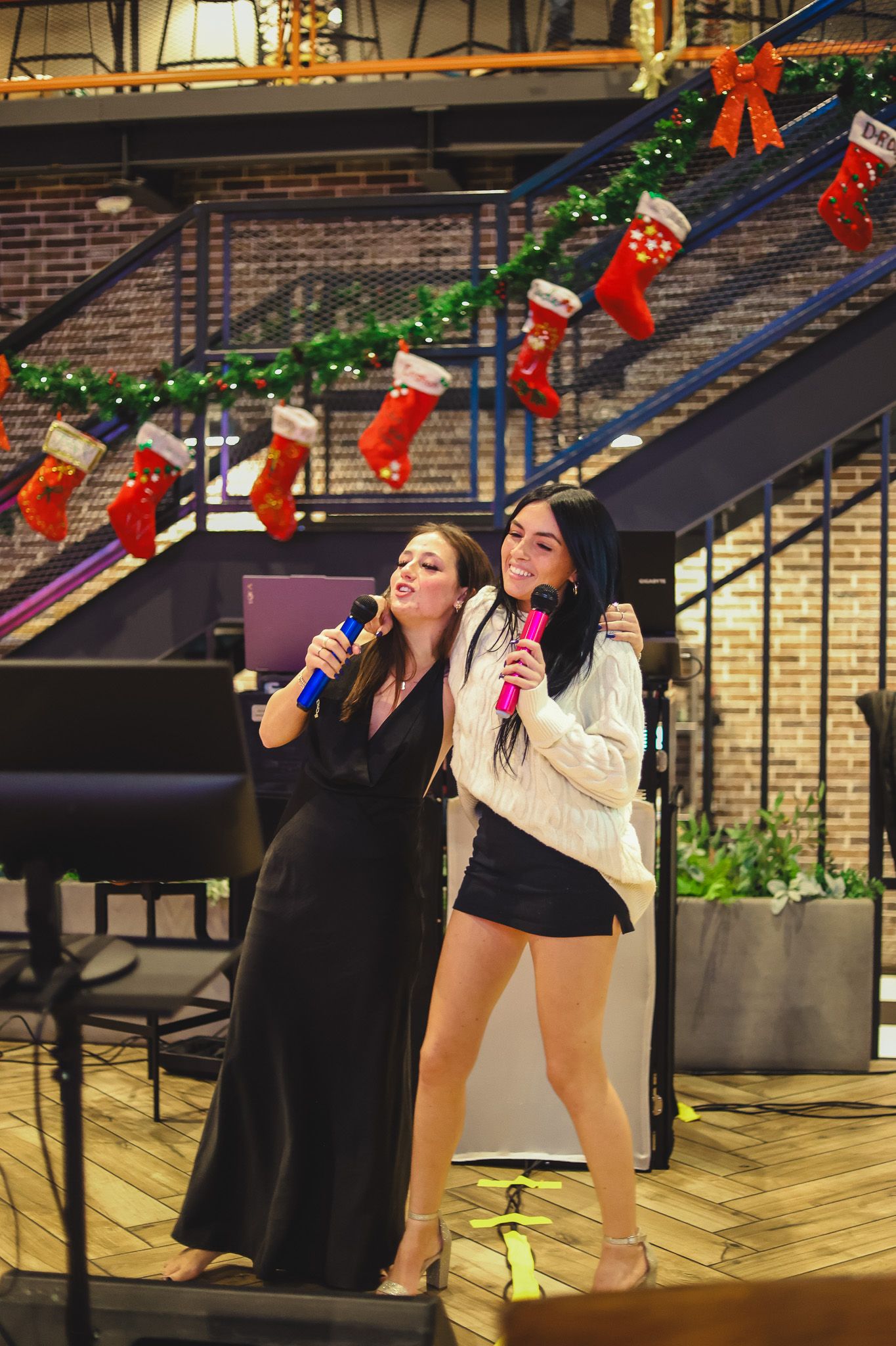Two women smiling and singing karaoke together on stage with Christmas decorations in the background.