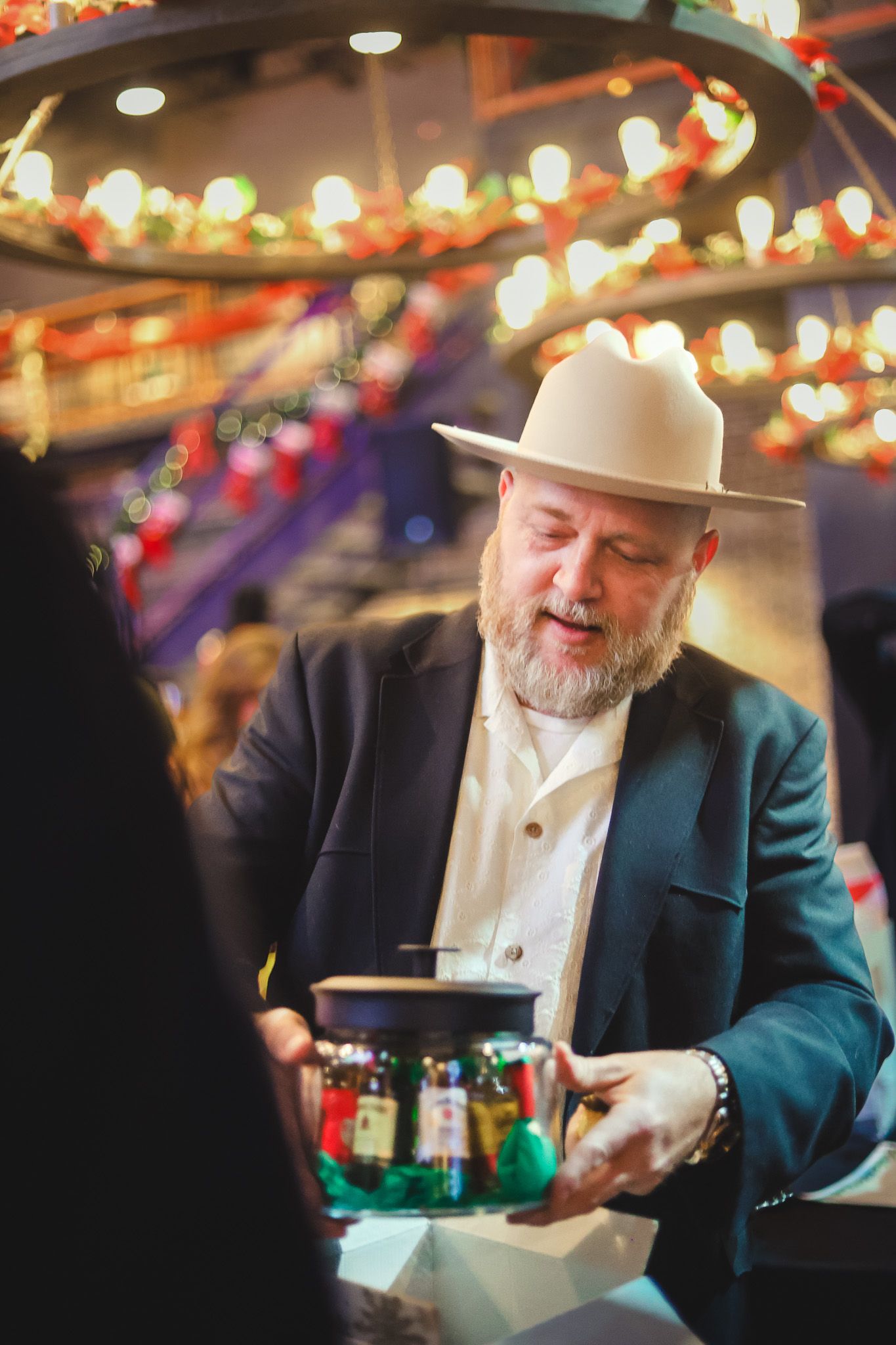 Bearded man in white hat and blazer holding a decorative jar at a festive event with colorful lights.