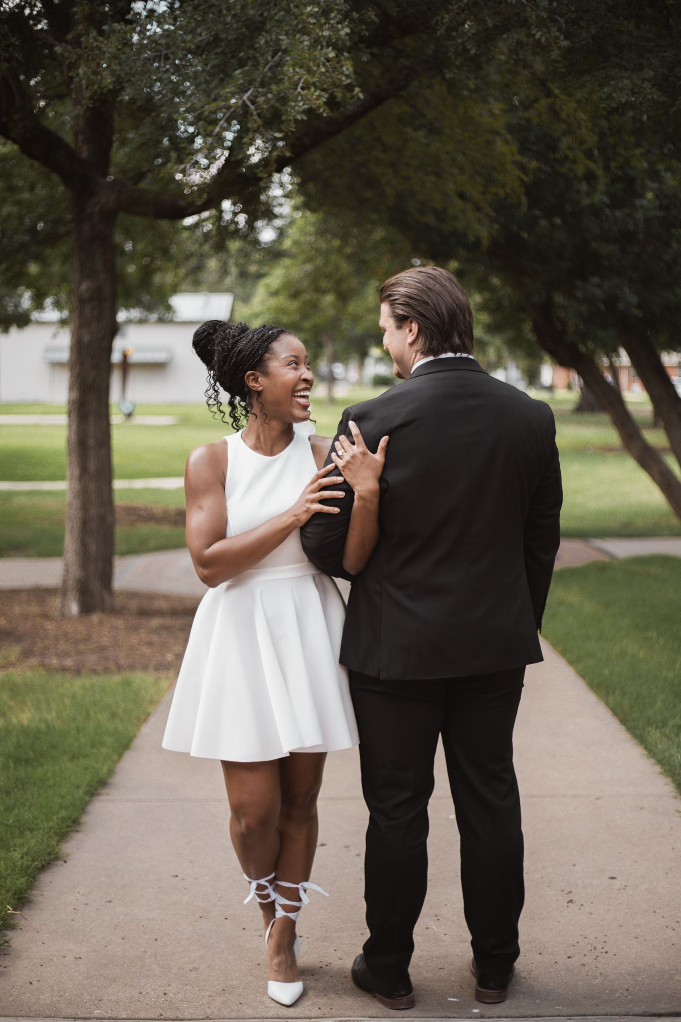 Interracial couple walking on a park path, woman in white dress smiling at man in black suit.