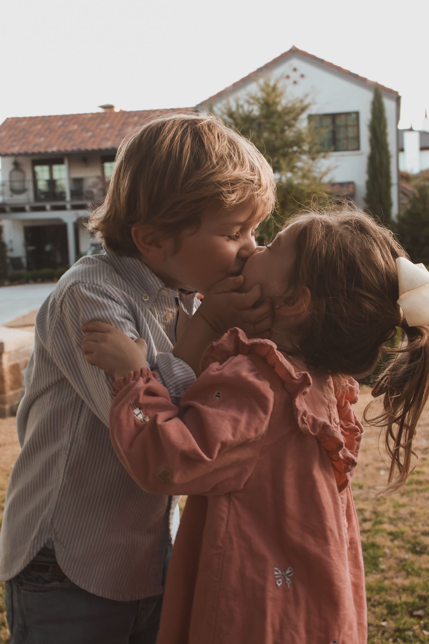 Two young children sharing a sweet kiss outdoors in front of a white building on a sunny day.