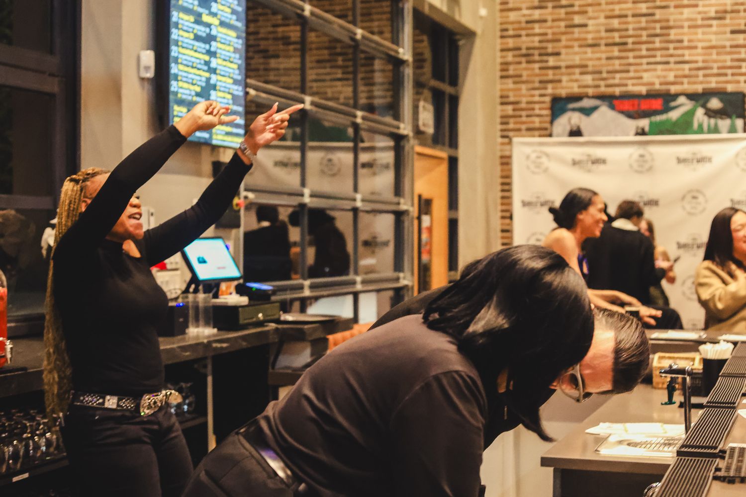 Women socializing at a bar counter in a warmly lit restaurant with brick walls and menu boards.