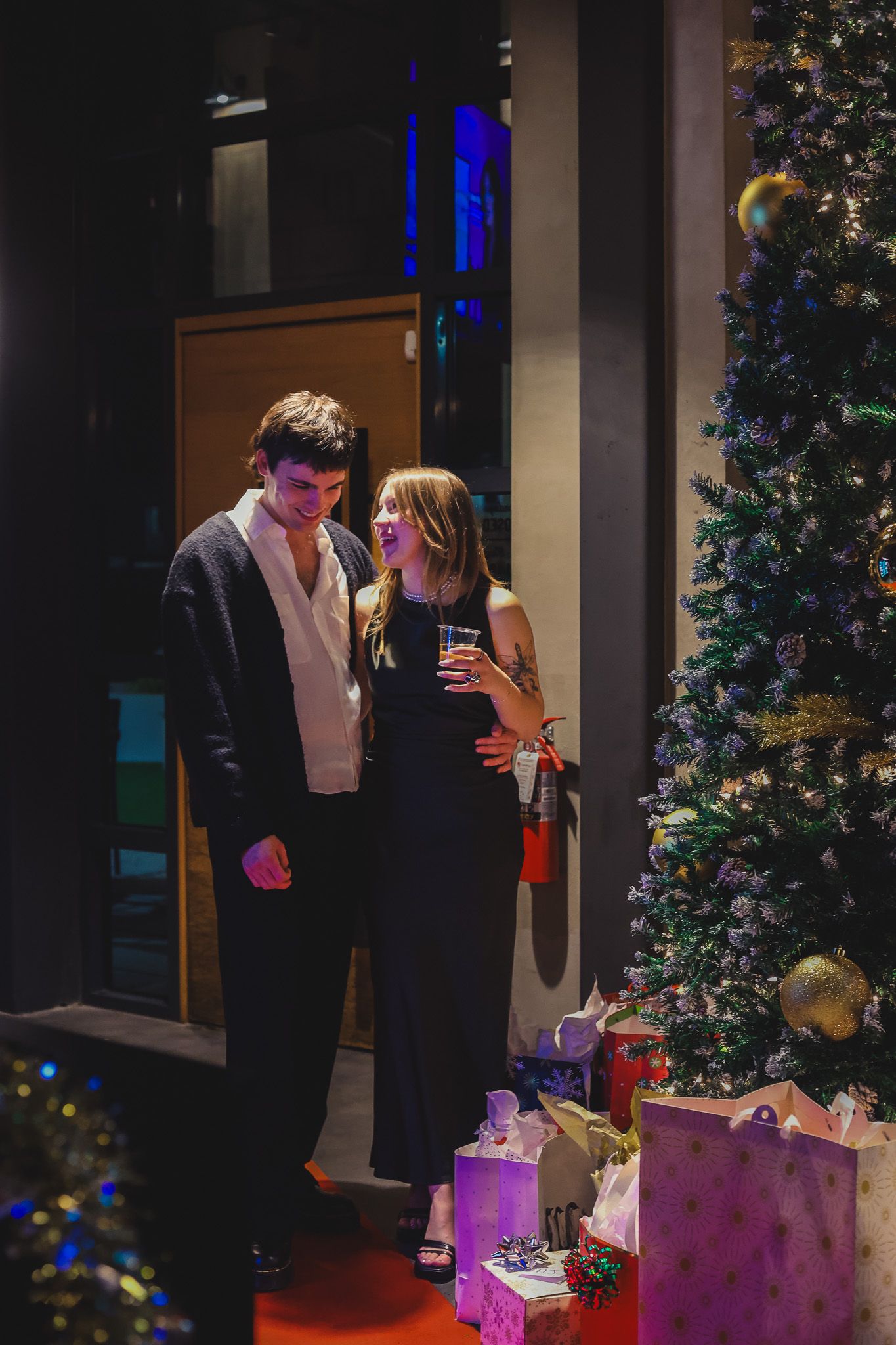 Couple in formal attire standing by a decorated Christmas tree with gifts at a holiday party.