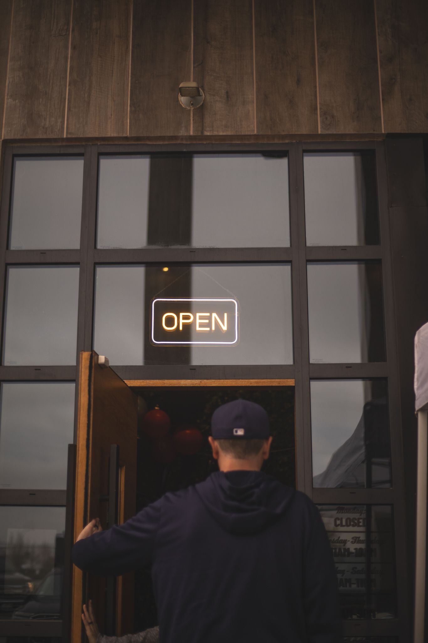 Man in baseball cap opening door of rustic wooden building with glowing neon OPEN sign.