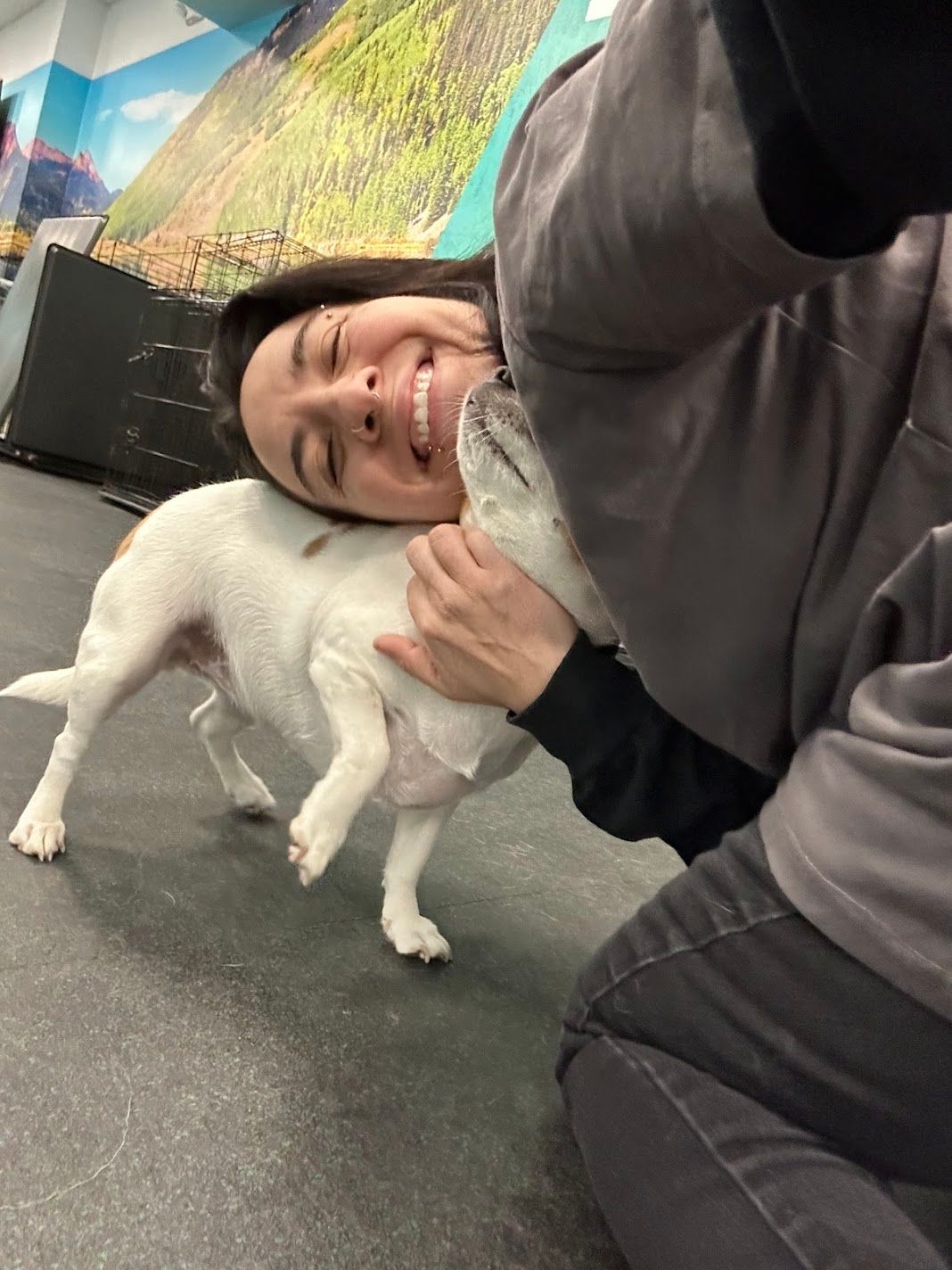 A joyful woman hugs a small white puppy on the floor of a colorful dog daycare center.