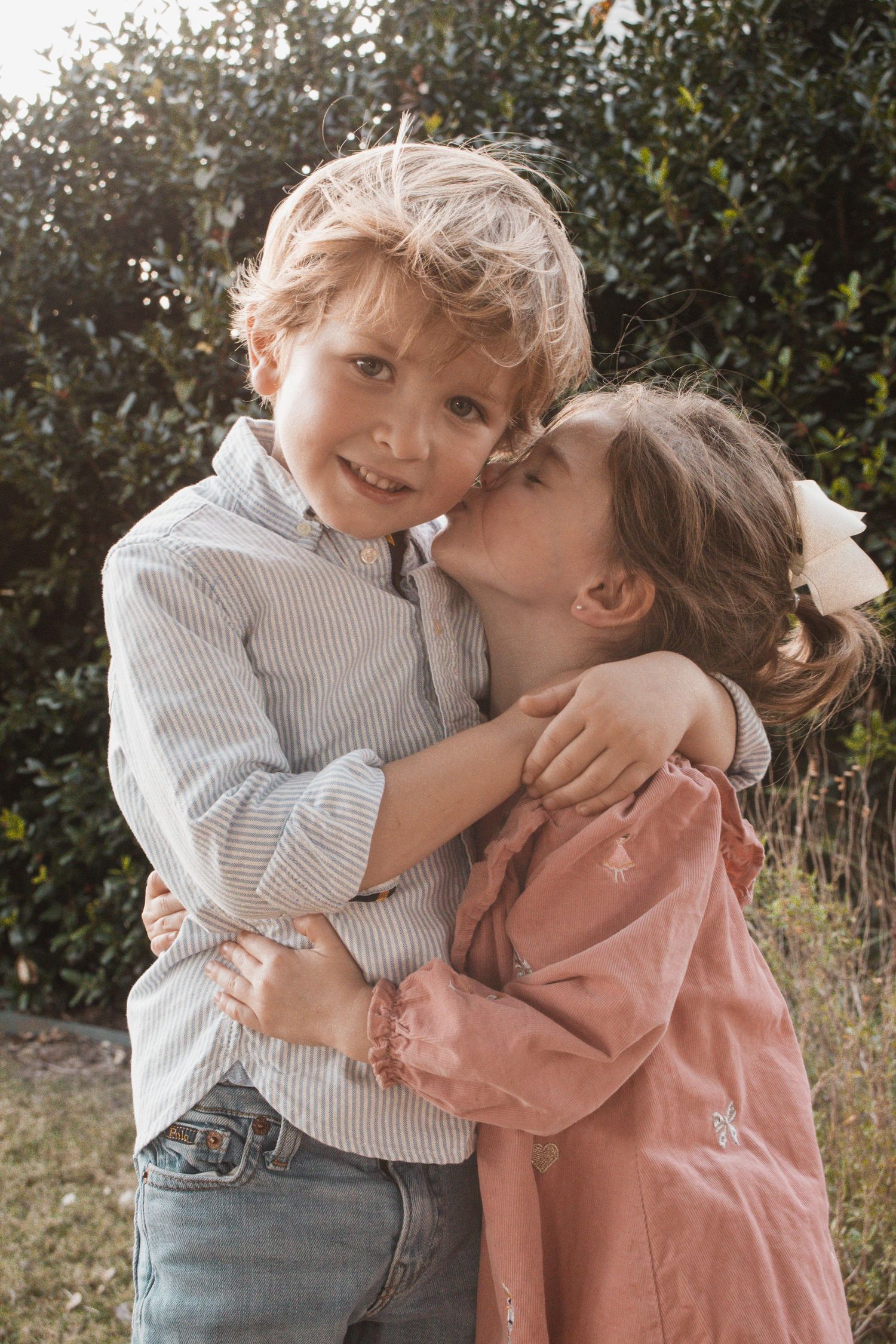 Two young children hugging outdoors, a smiling boy in a striped shirt and a girl in pink kissing his cheek.