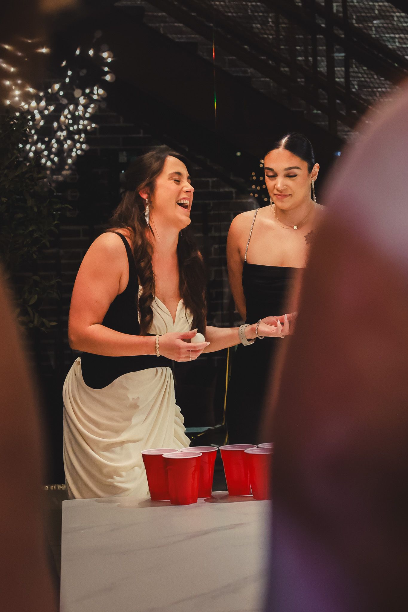 Two women laughing and playing beer pong with red solo cups at an outdoor evening party.