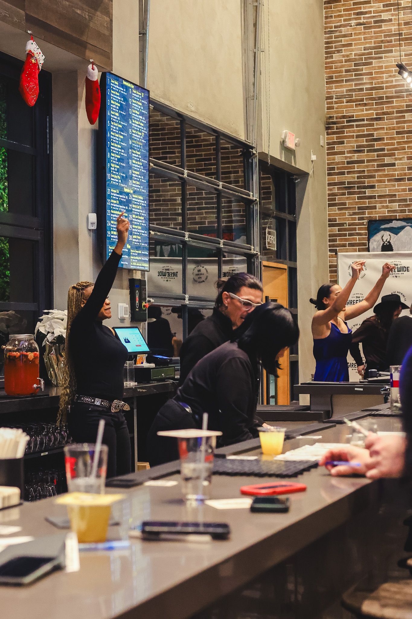 Busy bar scene with staff raising hands behind a counter, brick walls, menu board, and drinks on the surface.