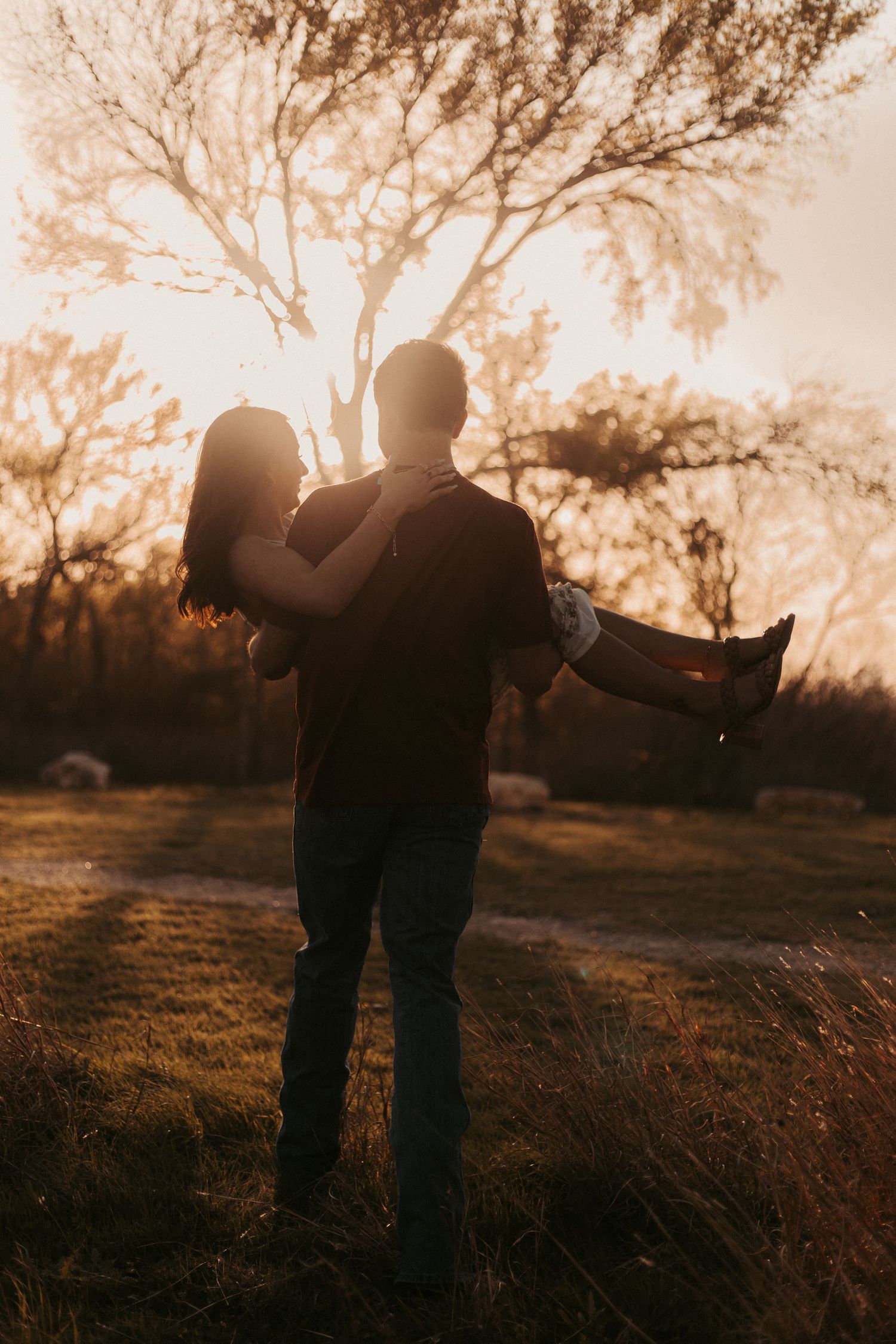 A couple shares a romantic sunset moment outdoors, man lifting woman in golden backlit light among trees.