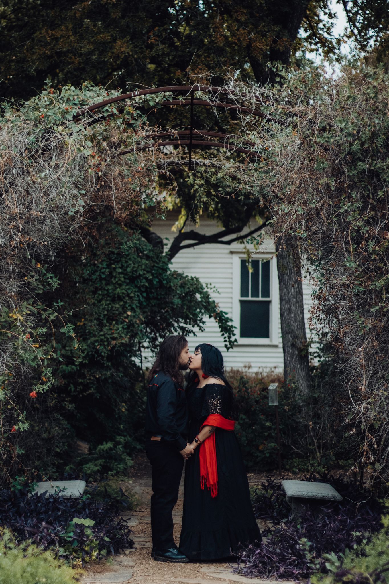Couple in black gothic attire sharing a kiss under a vine-covered arch in a lush garden setting.
