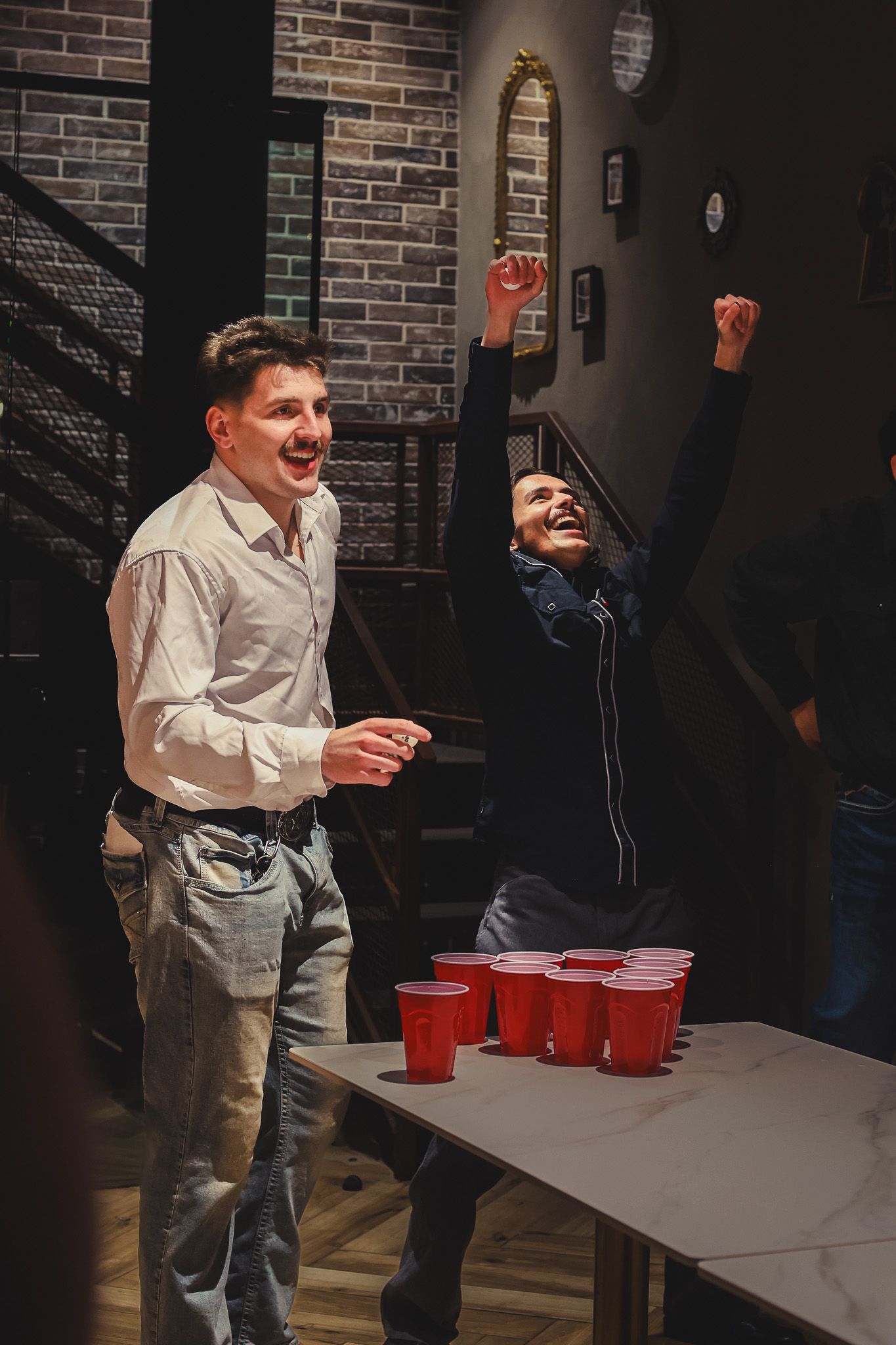 Two excited men celebrating while playing beer pong with red cups at an indoor party.