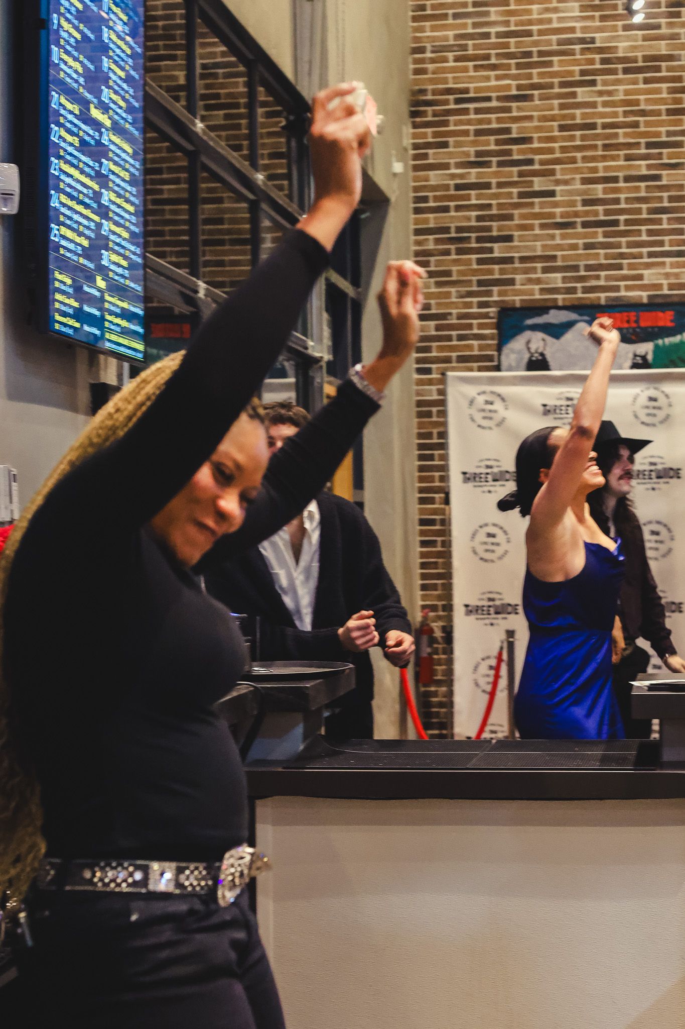 Two women raising hands at indoor event near brick wall with scoreboard display.