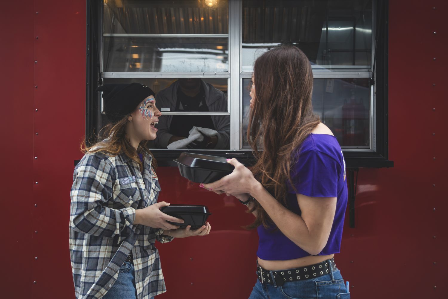 Two young women talking and exchanging items outside a red building with large windows.