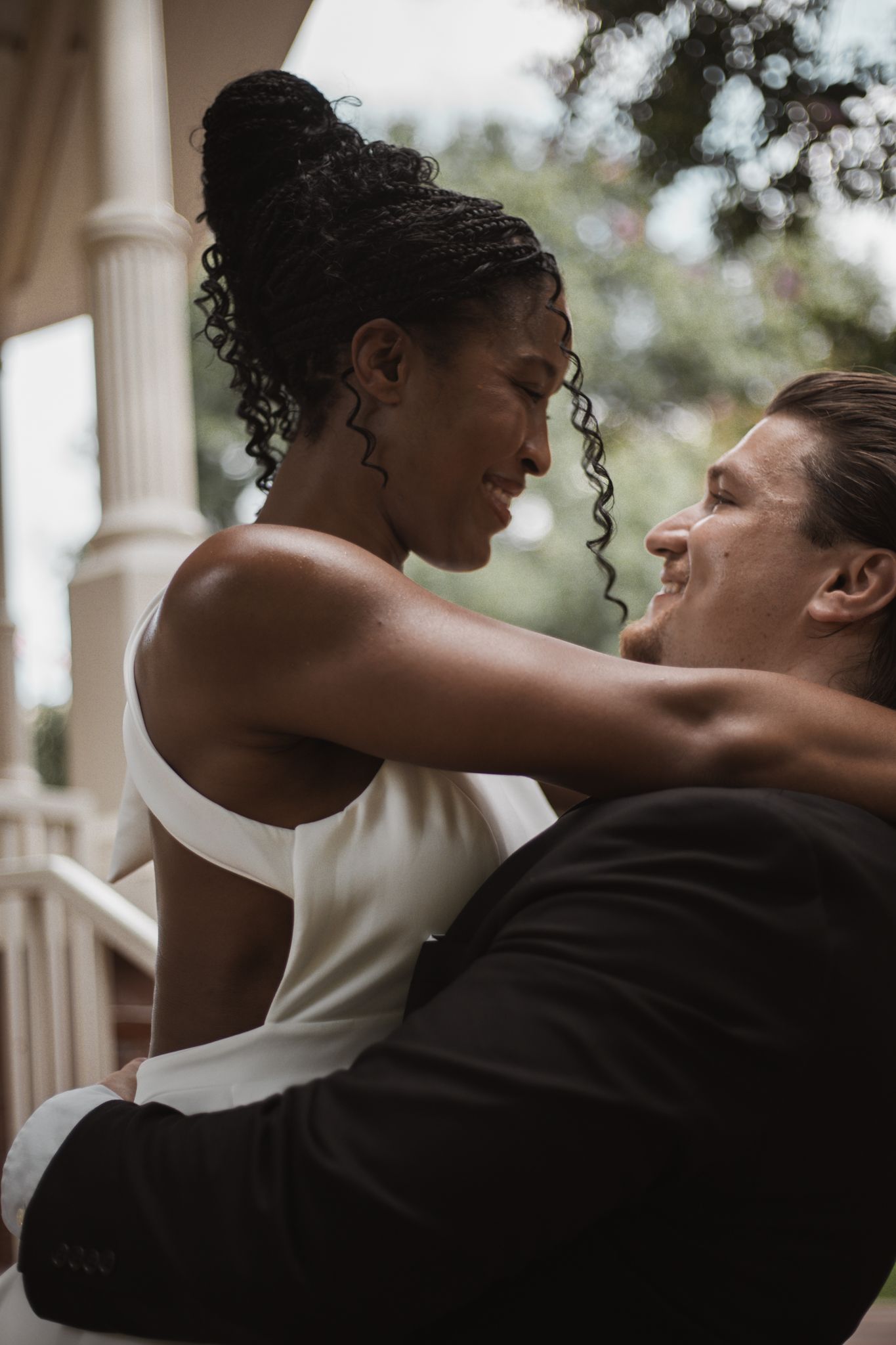Interracial couple sharing a romantic moment on a porch, bride in white dress smiling at groom in black suit.