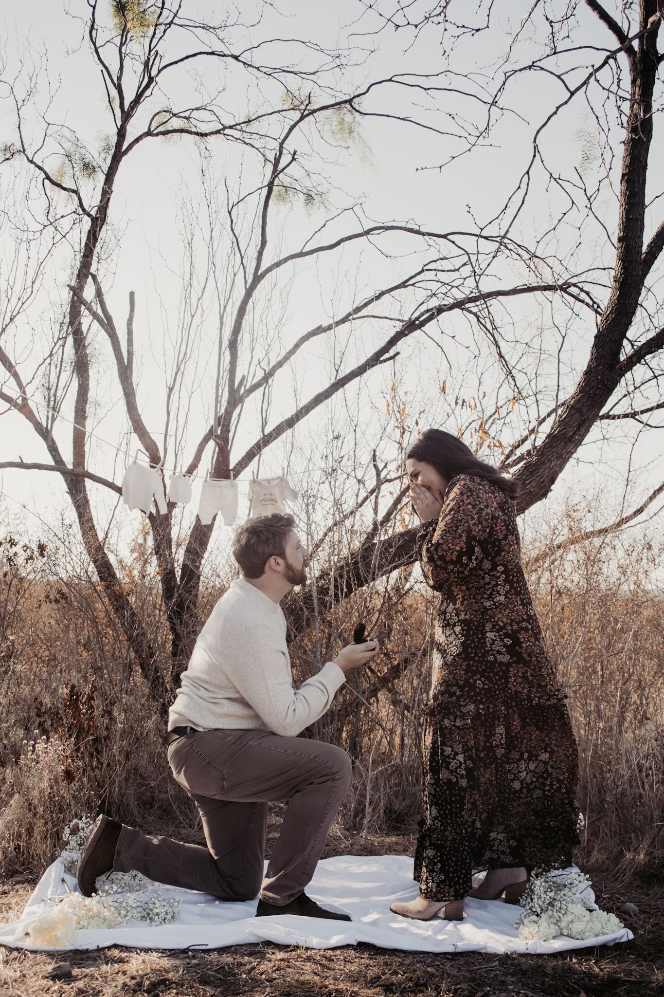 Man kneeling on white blanket proposing to woman in floral dress outdoors among bare trees in autumn.