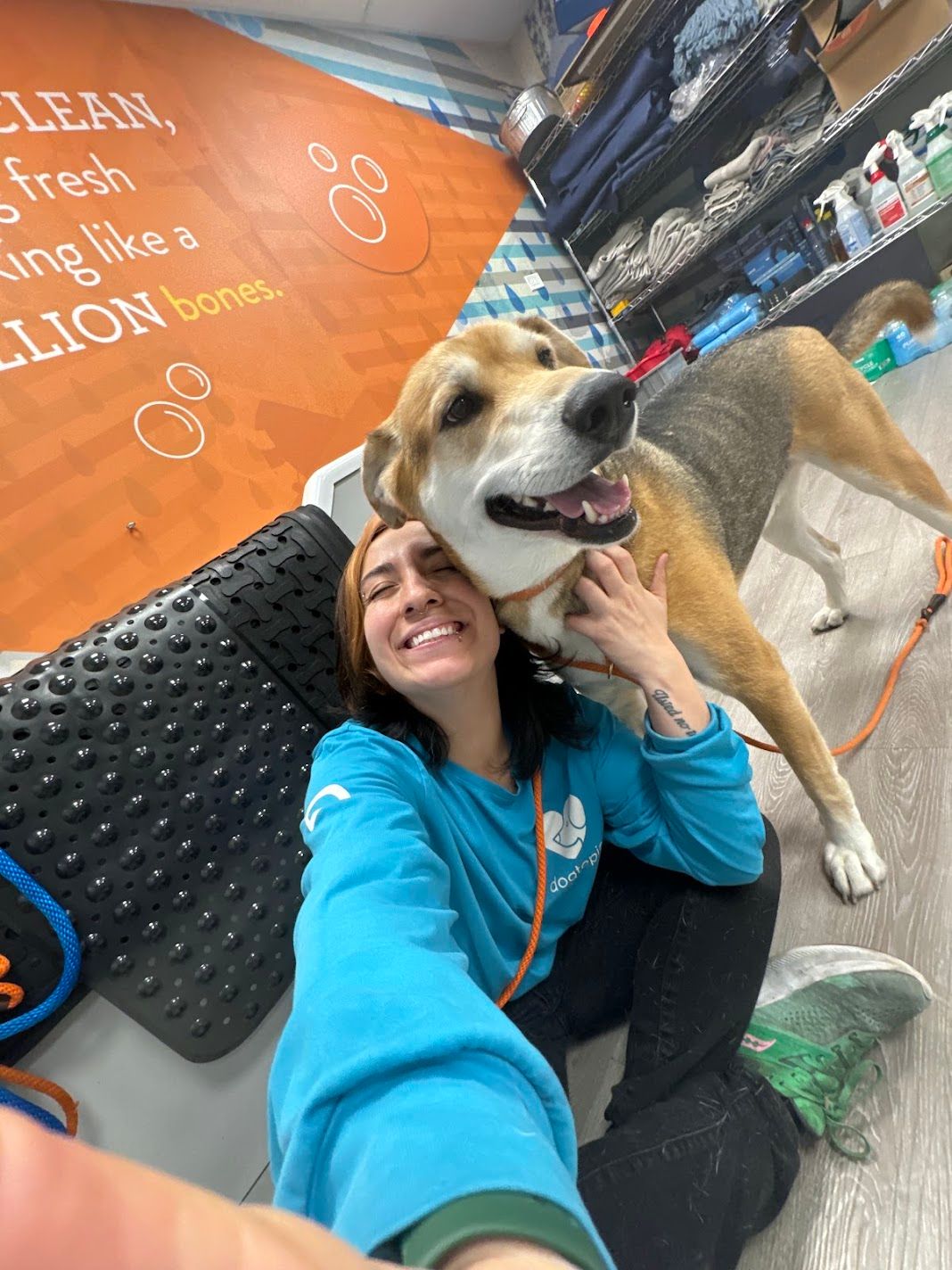Smiling woman in teal hoodie taking a selfie with a happy beagle mix dog at a pet grooming store.
