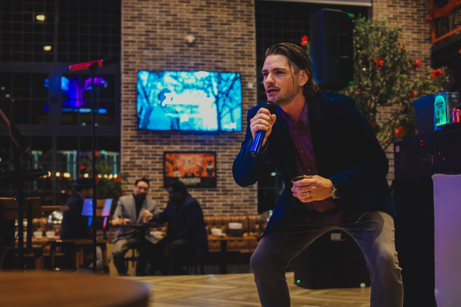 A man speaking into a microphone at an indoor venue with brick walls and ambient lighting during an event.