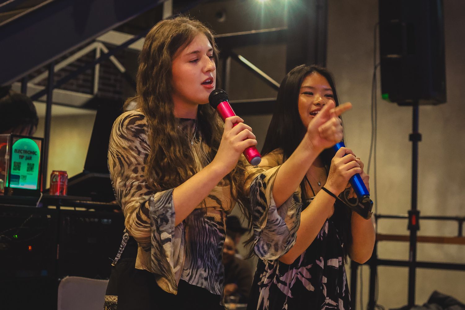 Two women singing karaoke together on stage, one holding a pink microphone and the other a blue microphone.