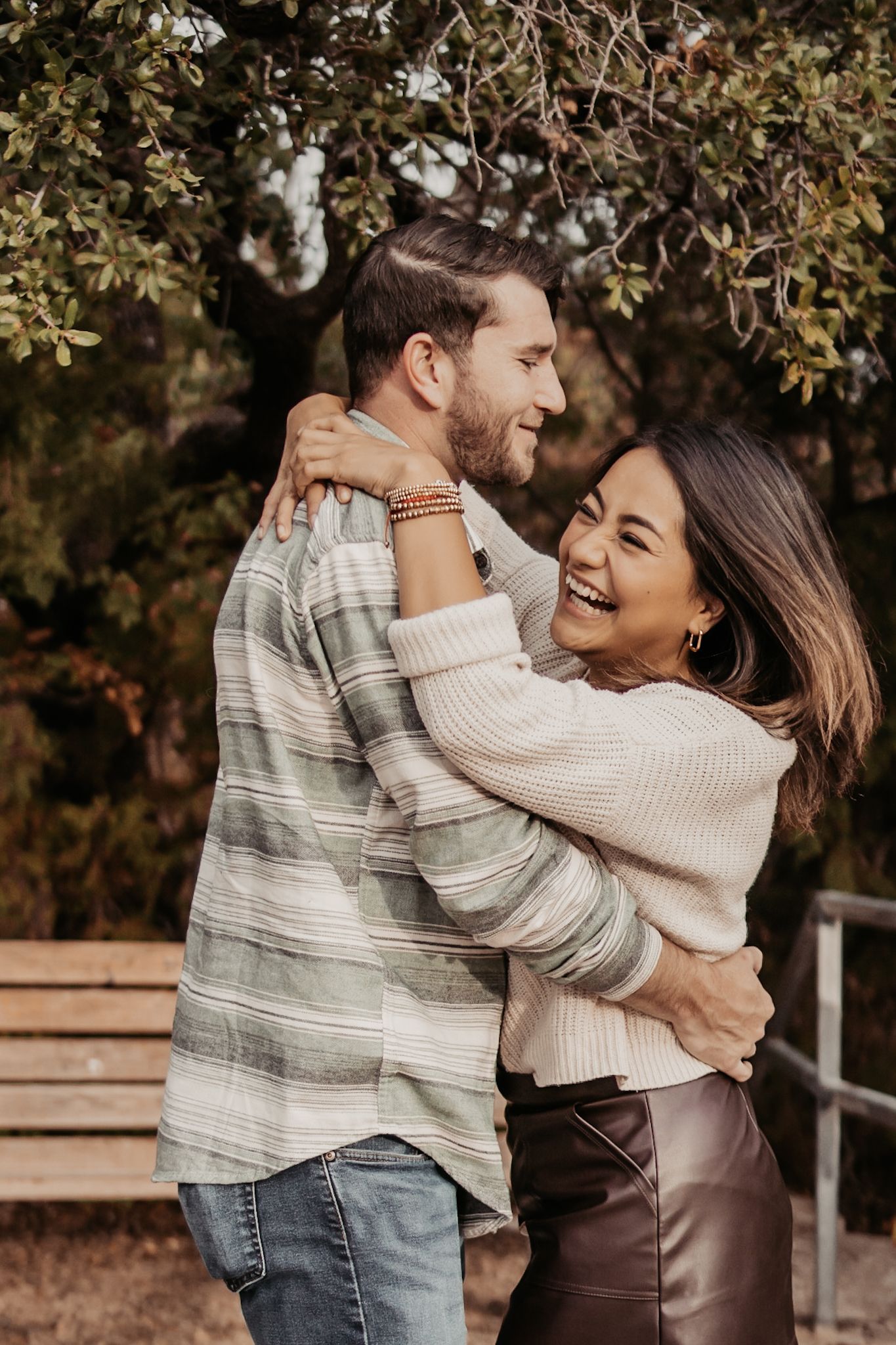 A joyful couple embraces and laughs together outdoors, with the woman in a cream sweater and leather skirt hugging a man in a striped shirt.