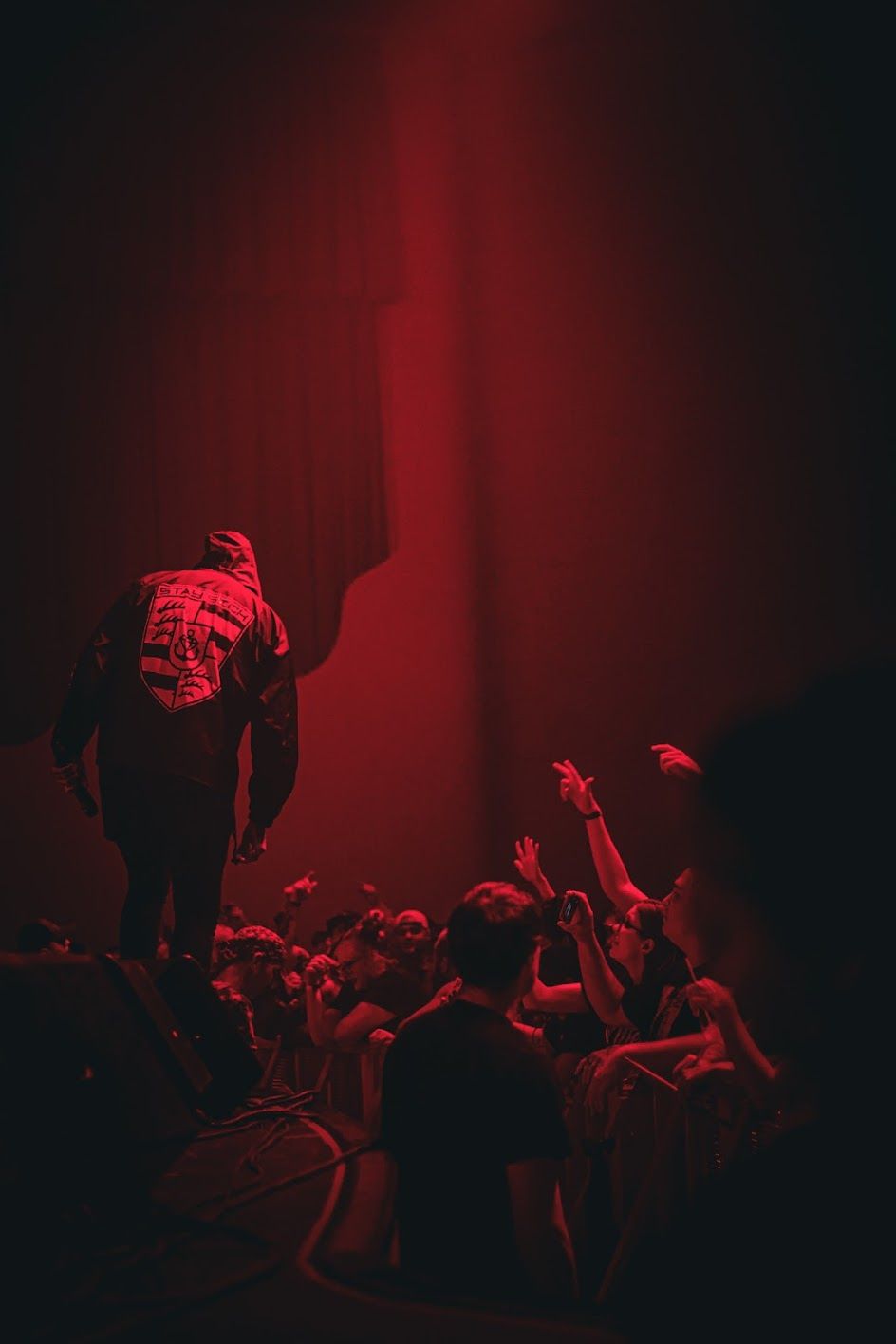 Concert performer standing above crowd bathed in dramatic red stage lighting at live show.