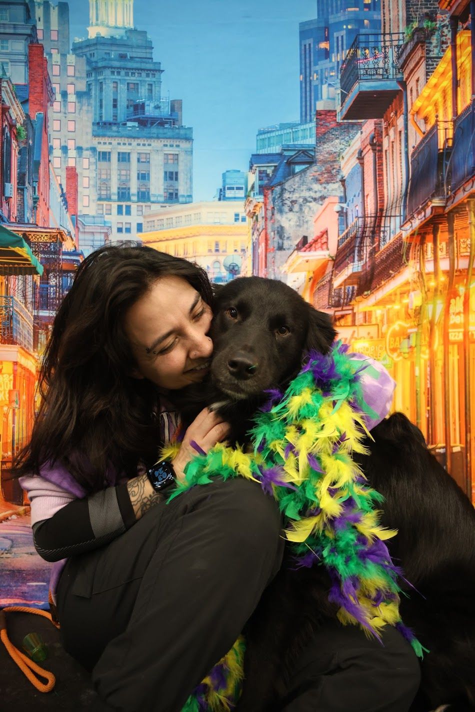 Young woman kissing a black dog wearing a Mardi Gras feather boa against a New Orleans backdrop.