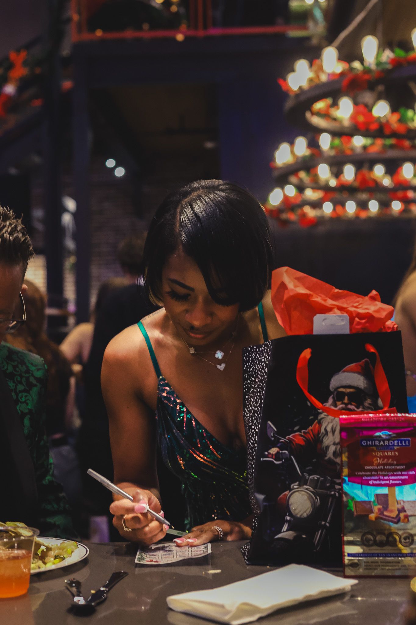 Woman in sequin dress writes notes beside a Ghirardelli gift bag at holiday party.