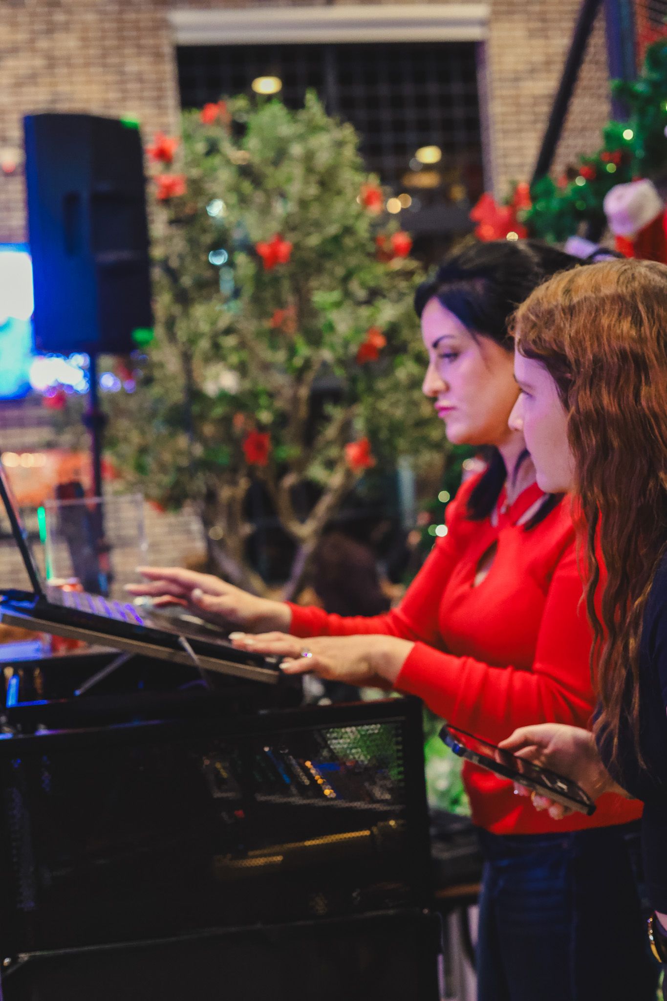 Two women in red tops playing piano together at a festive Christmas-decorated venue.