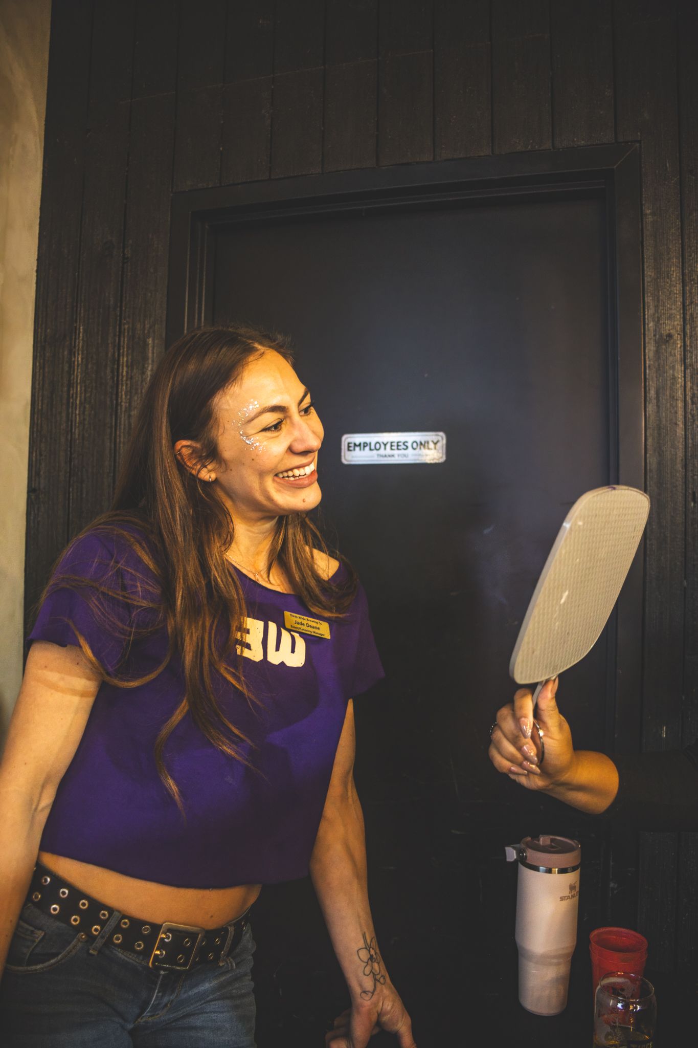 Smiling woman in purple crop top holding hand mirror near employees only door.