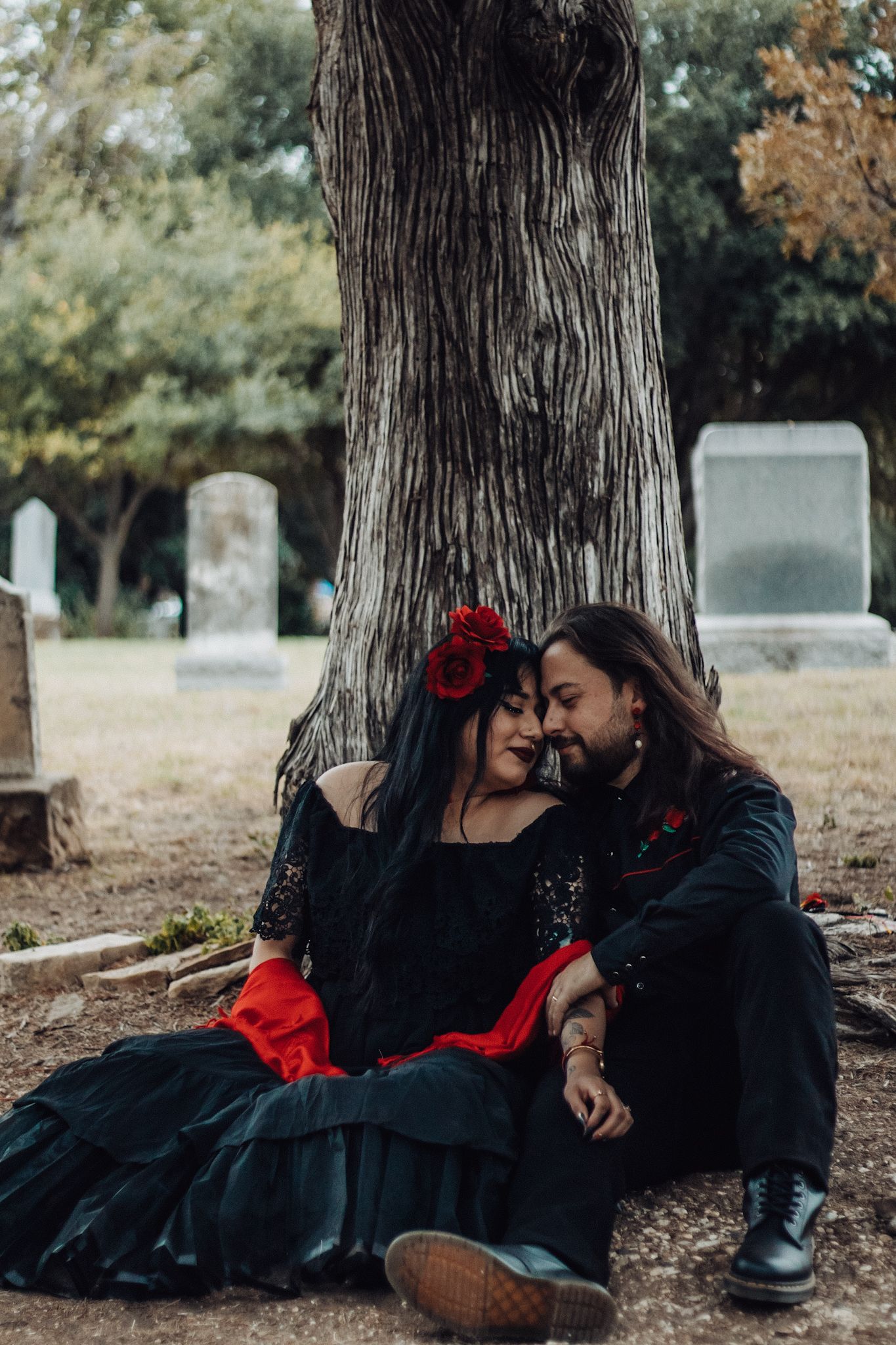 Gothic couple in black and red attire sitting romantically under a tree in a cemetery setting.