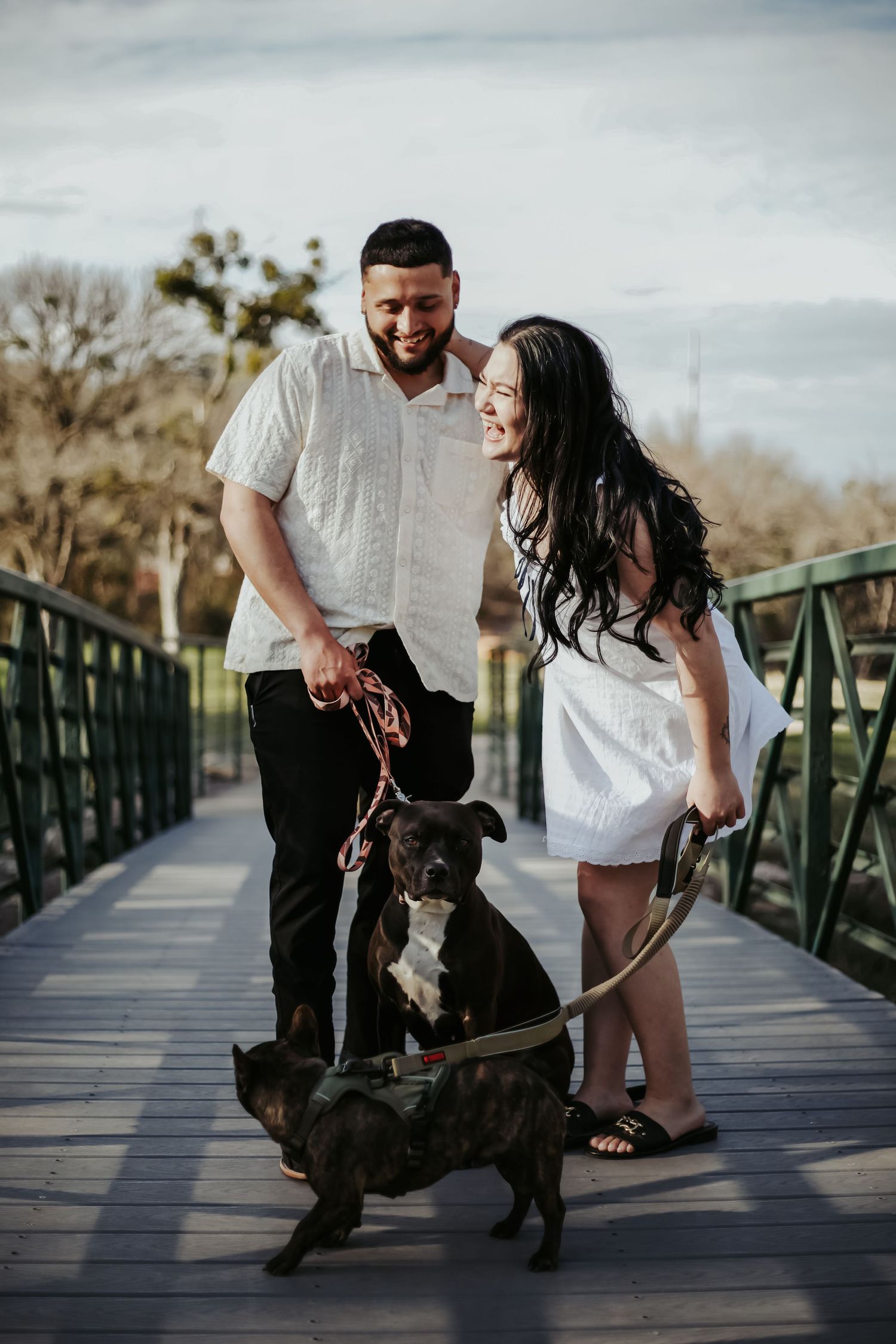 Smiling couple in white outfits walking their dark brindle Boxer dog on a wooden bridge outdoors.