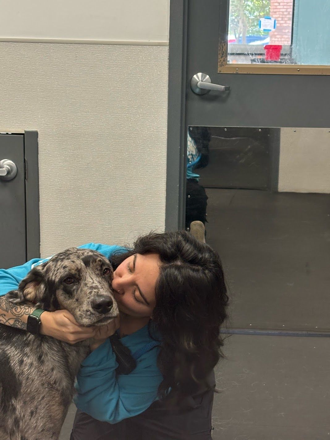 Woman in teal shirt hugging a gray spotted dog affectionately inside an animal shelter facility.