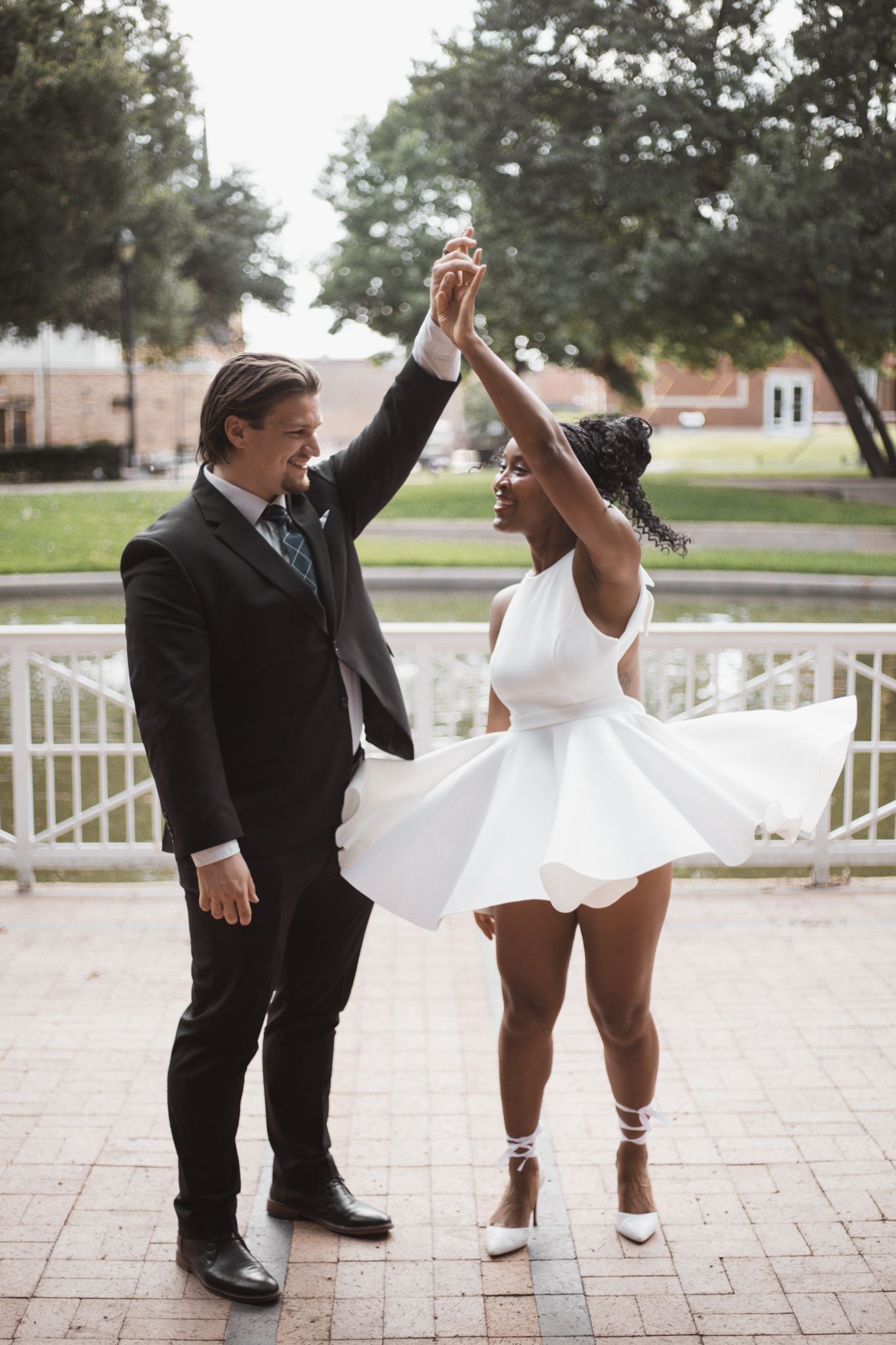 Interracial couple in wedding attire dancing joyfully outdoors near a fountain in a park.