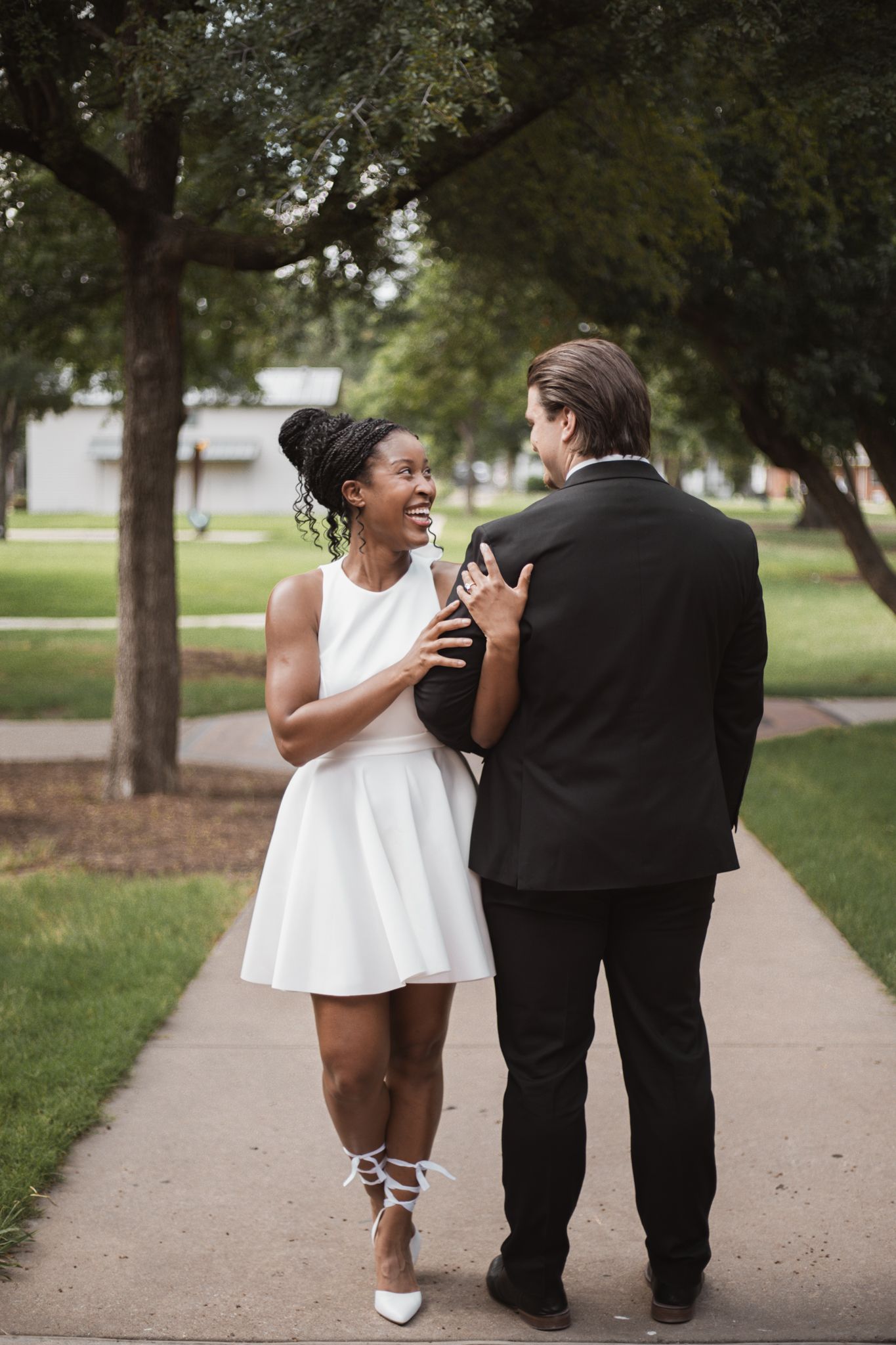 Joyful interracial couple walking together on a tree-lined sidewalk in wedding attire.