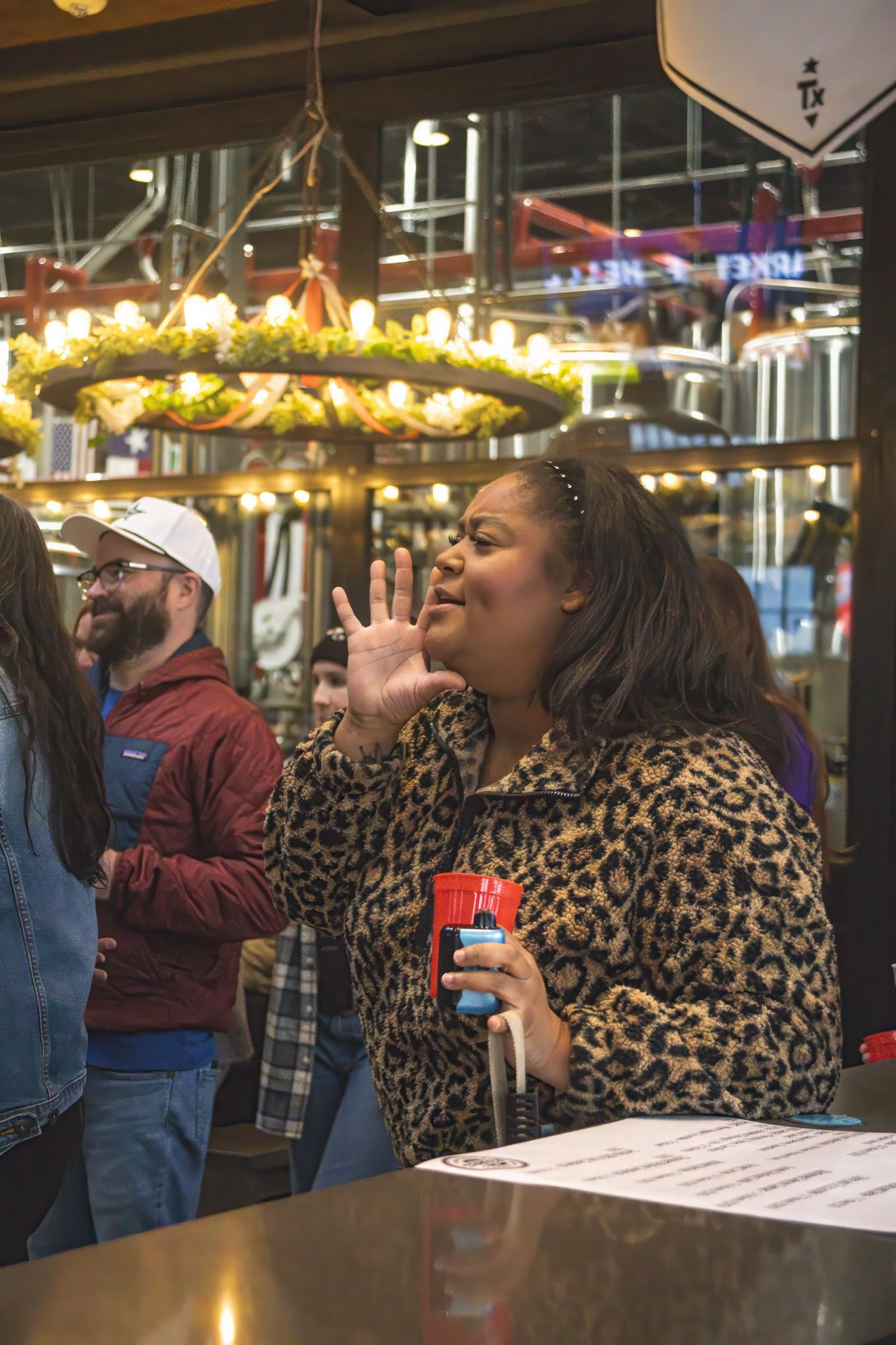 Woman in leopard print jacket raises hand holding red cup at lively brewery bar event.
